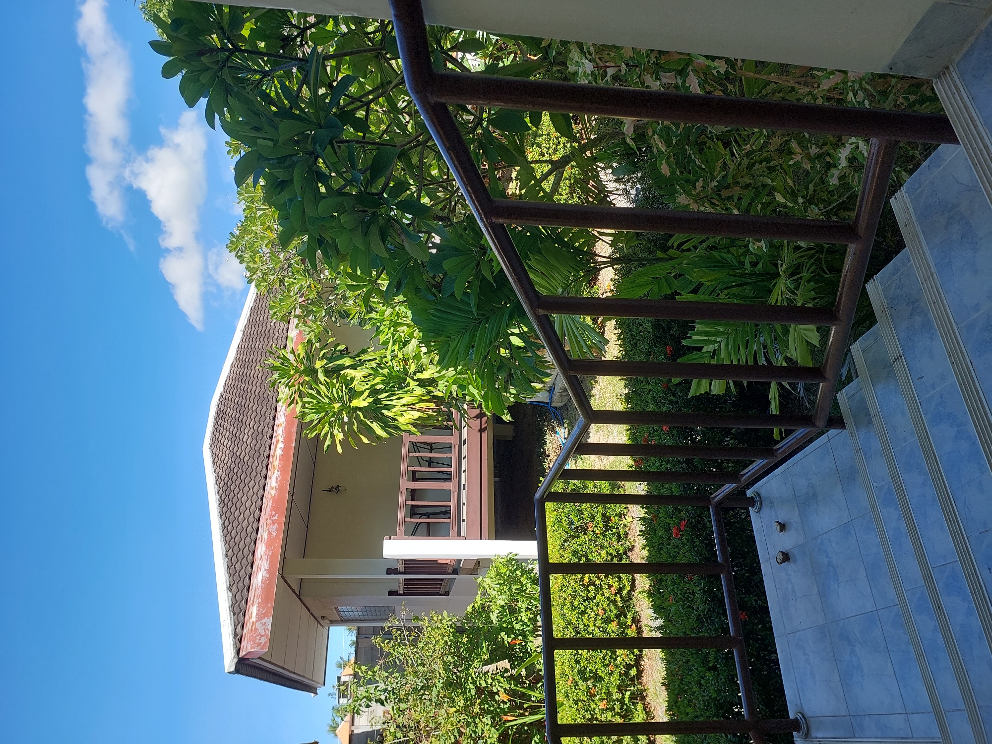 The image shows a view of a bungalow in Baan Mae Nam, Thailand, from a staircase. A brown railing with vertical supports is in the foreground, leading up a tiled staircase. The staircase landing at the top has two small, dark offerings placed on it. Beyond the railing, a small, single-story bungalow with a tiled brown roof and a light-colored facade is visible. A wooden porch with a brown railing extends from the bungalow. Lush green foliage, including palm trees and other tropical plants, surrounds the bungalow and the staircase, suggesting a tropical or subtropical climate. The sky is a clear blue with a few white clouds. The scene appears to be outdoors during the daytime.