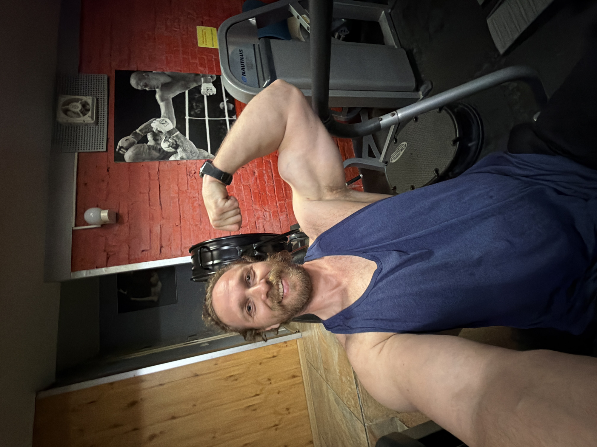 A man with a beard, wearing a navy blue tank top and a black smartwatch, flexes his bicep while smiling at the camera. He is in a gym with a Nautilus workout machine in the background. The wall behind him is red brick, adorned with black and white boxing photos and a ventilation fan. The flooring appears to be wooden or a wood-like material. The location in Saint Petersburg, Russia, is not discernible from the image.