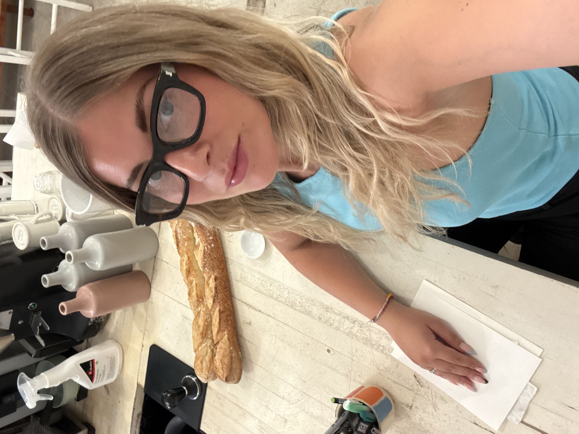 A woman with blonde hair and black-rimmed glasses is taking a selfie at a wooden table. She is wearing a blue tank top. In front of her, on the table, are two baguettes, a small white dish, a cup with pens and markers, and a stack of white paper. In the background, there are several white bottles and a spray bottle. The scene appears to be indoors, possibly in a cafe or restaurant, but there is no clear indication of a specific location in Paris, France.