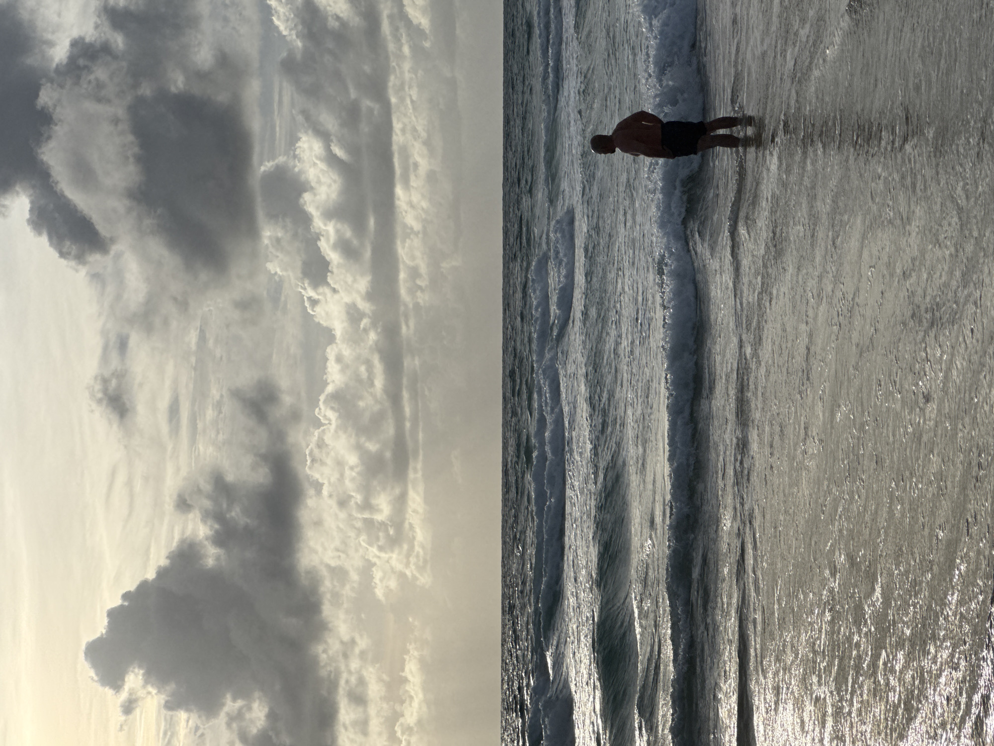 A solitary man stands waist-deep in the ocean at Karon Beach, Thailand, facing the sea. The sky above is filled with dramatic, grey clouds, with sunlight breaking through in places. The ocean is rough, with several waves breaking and washing ashore. The man is silhouetted against the bright water and sky, making his features indistinct. The overall mood of the image is one of solitude and contemplation, with the power of nature evident in the turbulent sea and sky.