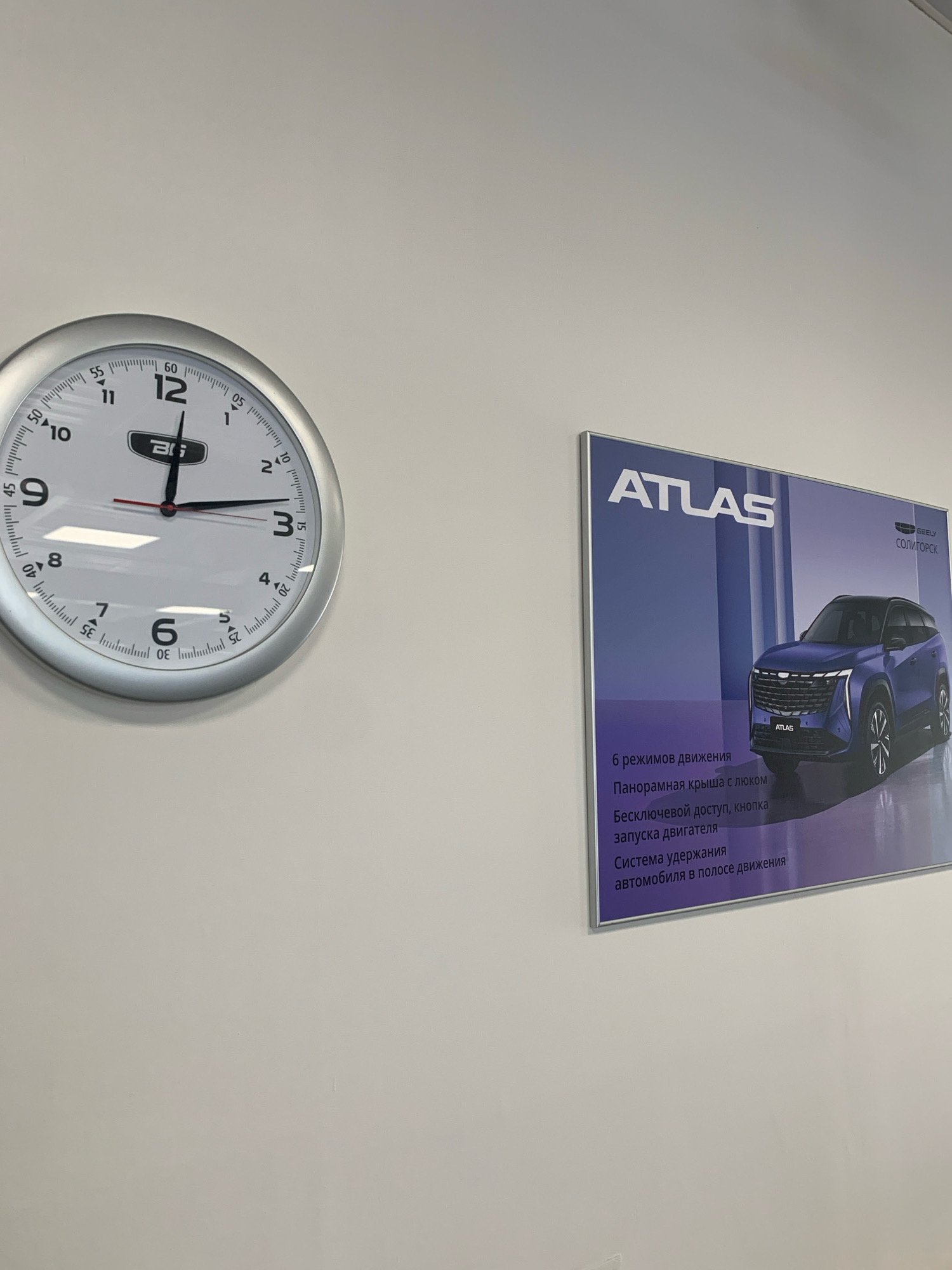 An indoor scene features a round analog clock on a light-colored wall, showing the time as approximately 12:42. The clock has a silver bezel and white face with black hour markers and numbers. A promotional poster for the Geely Atlas car is mounted on the wall to the right of the clock. The poster displays a blue Geely Atlas SUV and lists its features in Russian, including "6 driving modes," "Panoramic roof with sunroof," "Keyless access, engine start button," and "Lane keeping assist." The poster also has the Geely logo and the text "СОЛИГОРСК," indicating its location in Soligorsk, Belarus. The environment appears to be a clean, well-lit interior, possibly a car dealership or showroom. There are no people visible in the image. The clock face reflects some overhead lighting.