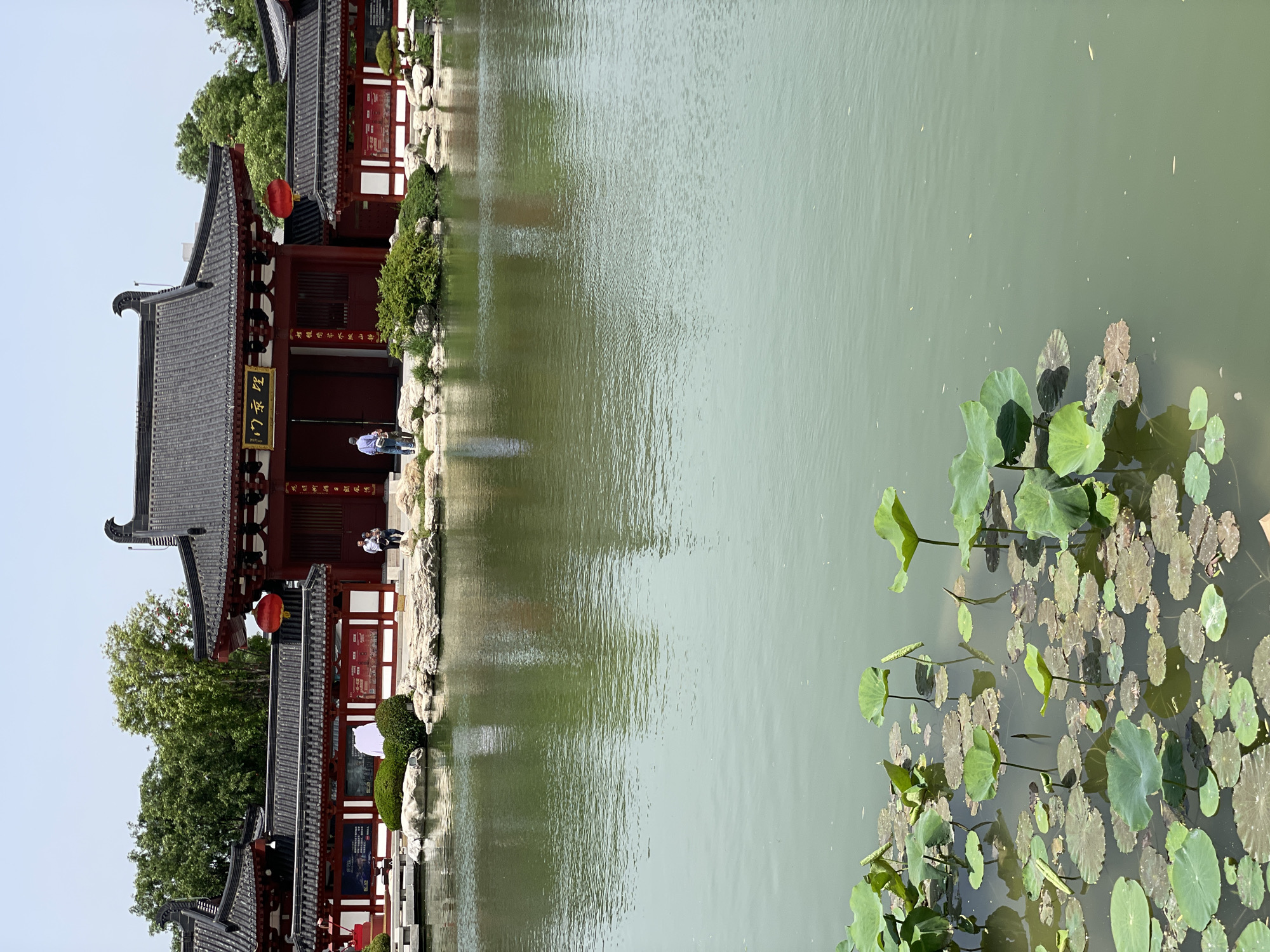 The image shows a traditional Chinese building, possibly a temple or pavilion, situated beside a body of water. The architecture features dark tiled roofs, red painted wooden structures, and decorative details such as lanterns. A stone pathway runs along the edge of the water, with people standing on it. The water reflects the building and sky. Lotus pads with green leaves float on the water's surface. The picture location is in Lintong District, China.