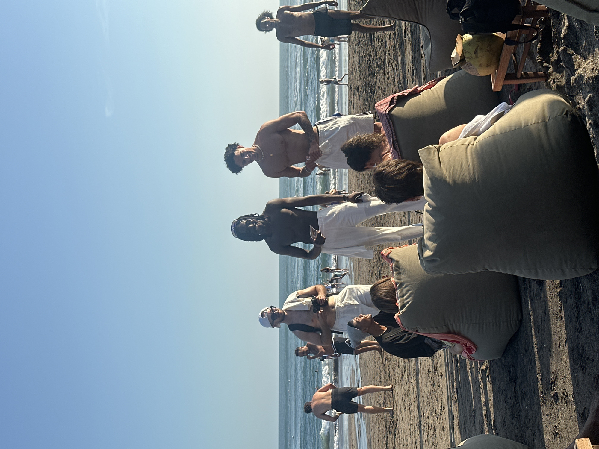 The image shows a group of people on a sandy beach, likely in North Kuta, Indonesia, on a clear, sunny day. The ocean with gentle waves is visible in the background, and the sky is a bright, clear blue.
In the foreground, several people are seated on large, light-colored beanbag chairs. One person in a black shirt is facing away from the camera, and another person, partially visible, is also seated. To the right, a green coconut with a straw rests on a small wooden stool. A black backpack is placed next to the stool.
Further back on the beach, three men stand talking. The man on the left, with dark skin and dreadlocks, is wearing white pants. The man in the center, with lighter skin, is wearing white shorts and a white cap. He is holding a phone and has a towel draped over his shoulder. The man on the right, with dark skin and curly hair, is wearing dark shorts.
Other people can be seen in the middle ground, some walking and some in the water. The overall atmosphere appears relaxed and social, characteristic of a beach day.