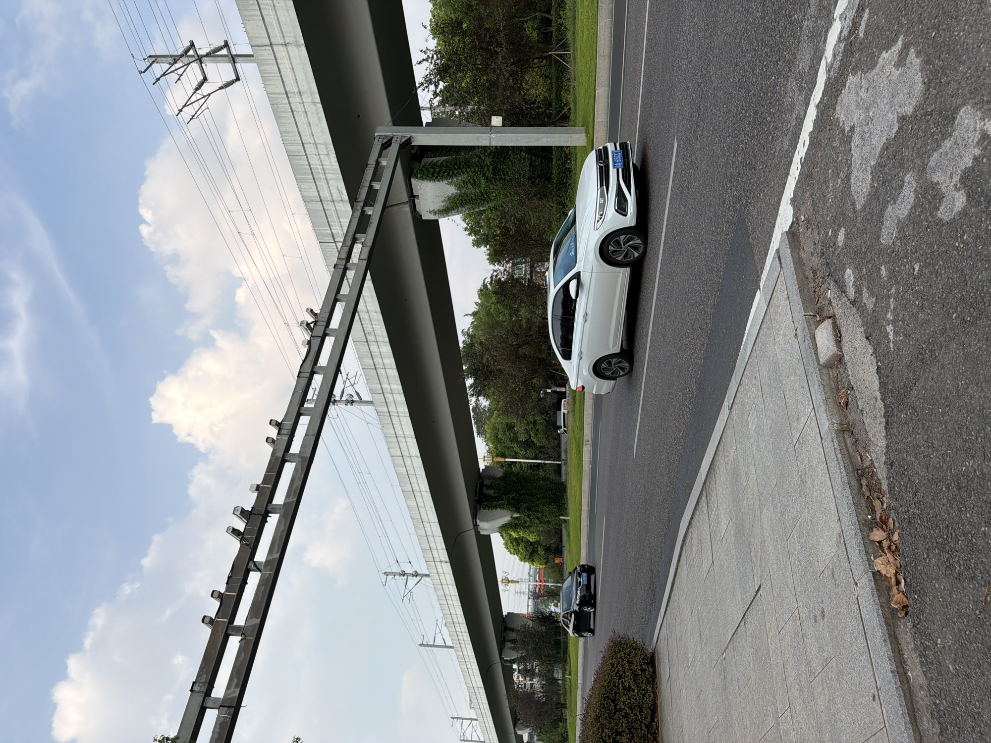 In the city of Jiangjin, China, on a paved road under an elevated structure that carries railway tracks and power lines, a white Volkswagen sedan with license plate "A 650CJ" drives on the right lane of the road. A black sedan is visible driving in the left lane. The sky is partly cloudy with a pale blue hue. The road is bordered by a sidewalk on the left, and a grassy median with trees on the right. The elevated structure is supported by concrete pillars covered in green ivy. A few fallen leaves are scattered on the sidewalk and along the curb. The overall scene depicts a typical urban environment with transportation infrastructure and natural elements.