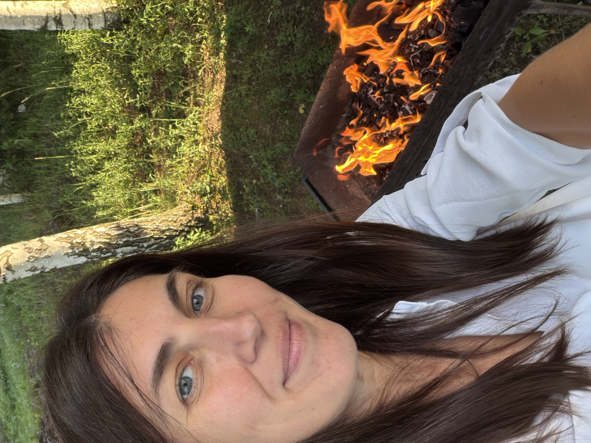A medium shot captured outdoors in Dubrova, Russia, features a woman in a white shirt posing for a selfie. Her brown hair frames her face, and her blue eyes look directly at the camera. She has a slight smile. Behind her, a metal grill with bright orange flames and glowing coals is visible. The background consists of lush green grass and trees, suggesting a natural or park-like setting. The lighting indicates it is daytime, with sunlight illuminating the scene.