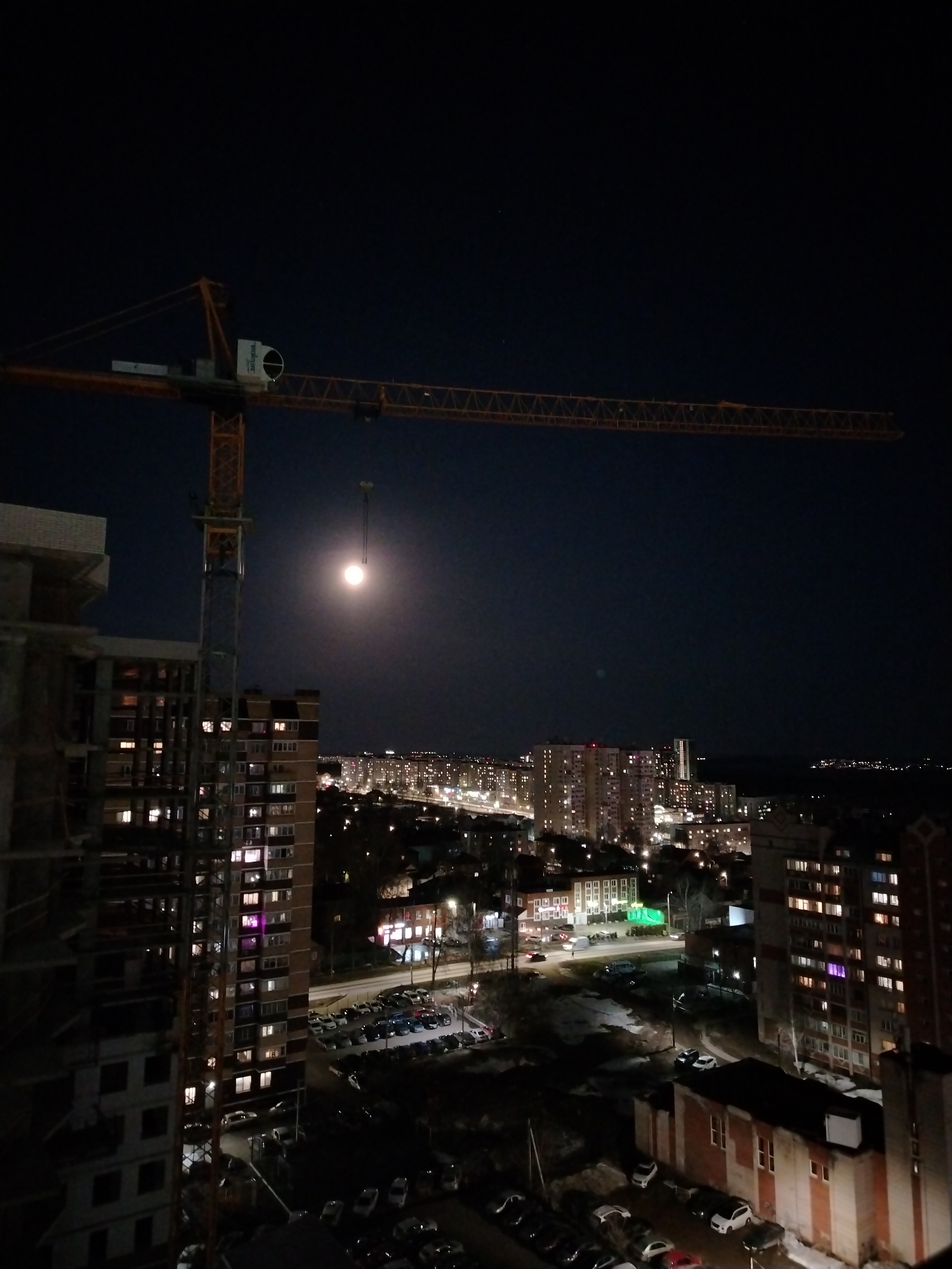 The image depicts a nighttime view of a city, likely Izhevsk, Russia, due to the distinct urban landscape and architectural styles. Dominating the foreground is a construction site with a large yellow crane, partially obscuring the full moon. A high-rise building under construction is visible to the left of the crane. The middle ground consists of residential buildings with illuminated windows, suggesting activity. Streetlights and car headlights illuminate the streets. In the background, a cityscape stretches into the distance, with numerous buildings and lights defining the urban skyline. The presence of parked cars and the general layout of the city contribute to the urban environment.