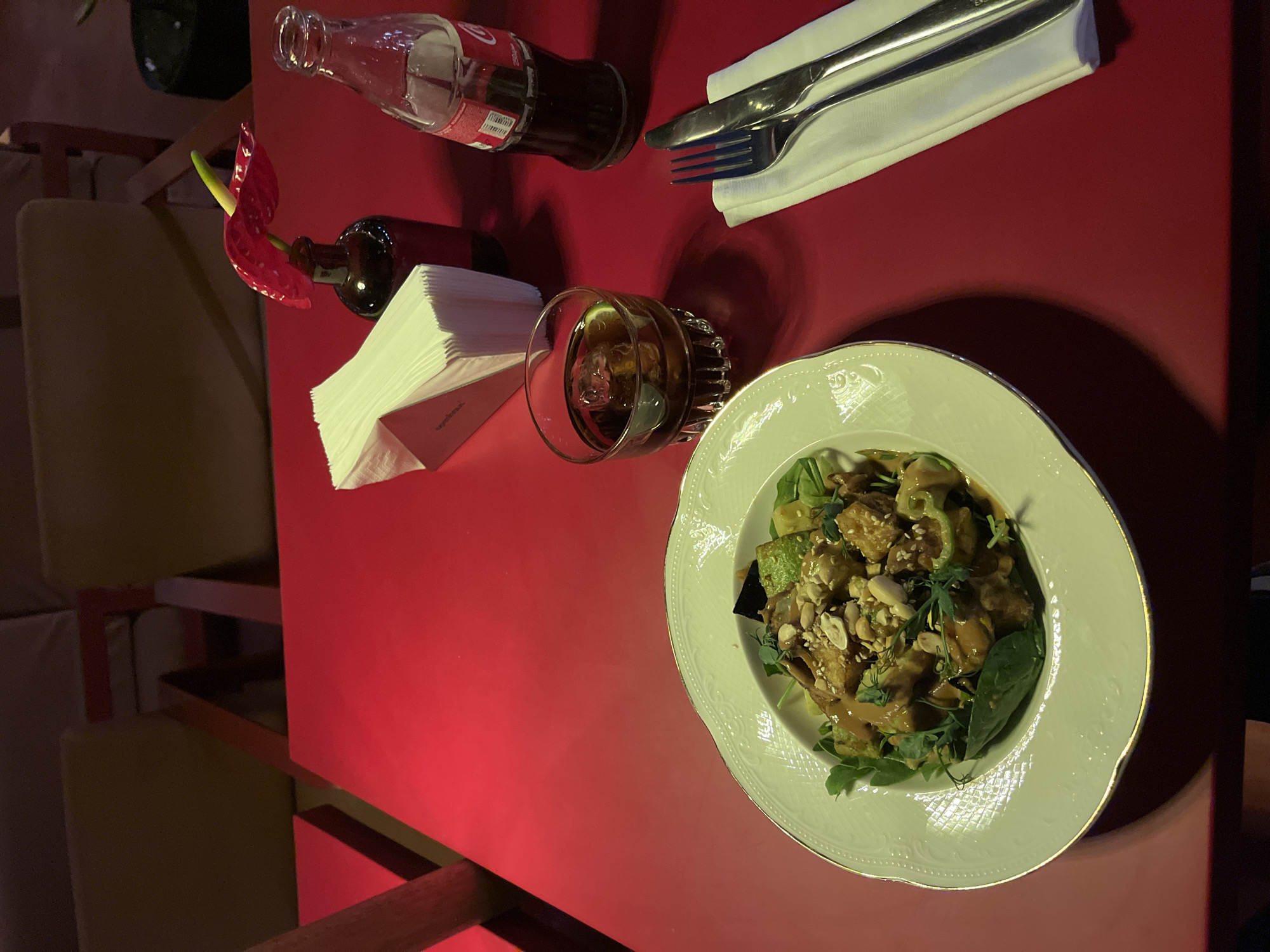 The image depicts a table setting in what appears to be a restaurant or cafe. The table is covered with a red surface. A plate containing a salad with visible peanuts and leafy greens sits in the lower right quadrant. A glass of liquid with ice cubes is placed near a stack of napkins and a small red-colored flower. A bottle of Coca-Cola and a set of utensils wrapped in a napkin are placed towards the upper left. The presence of a wooden frame suggests the setting is indoors, possibly in Yerevan, Armenia, based on the provided location information.