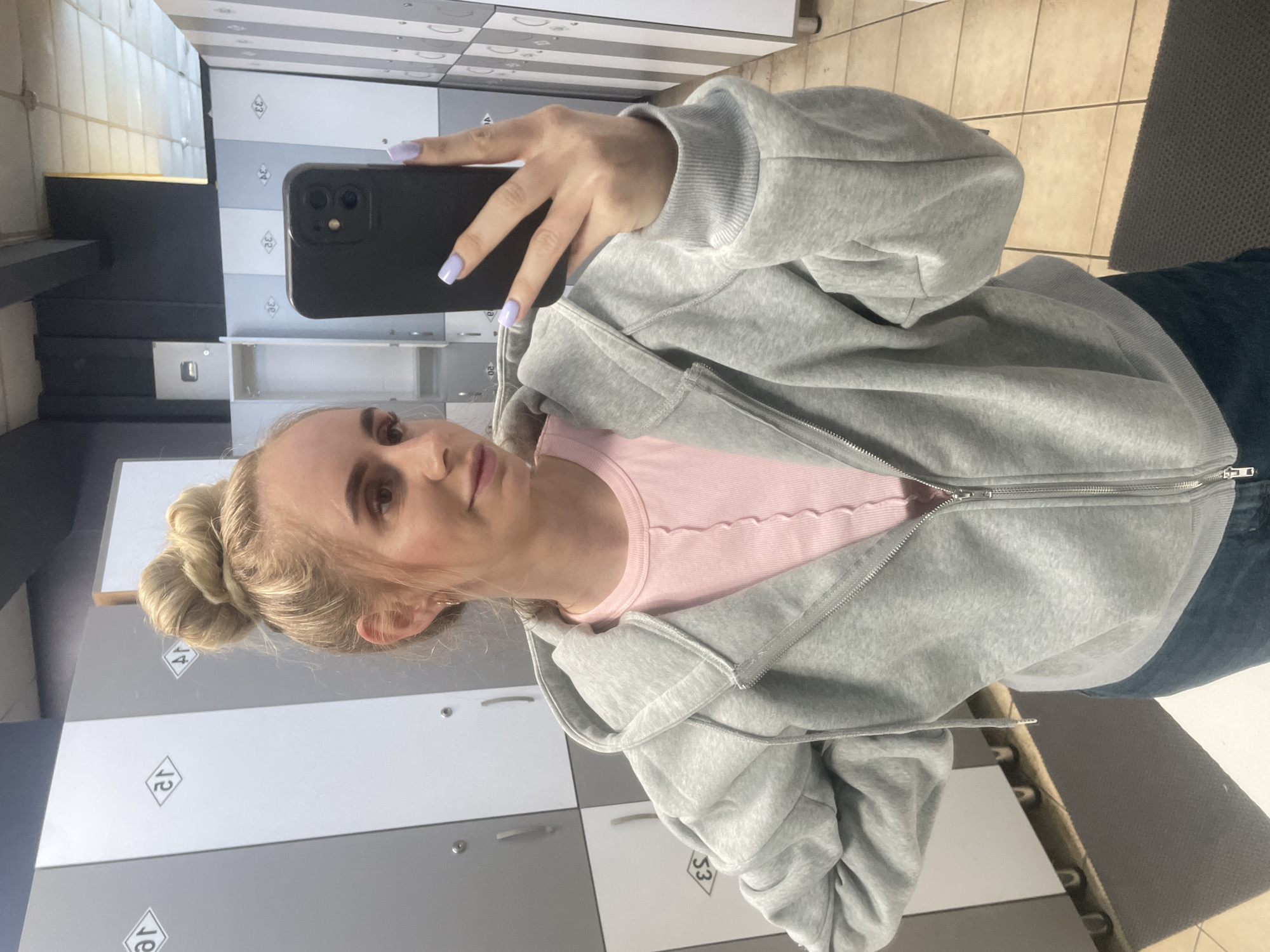 A young woman is taking a mirror selfie in what appears to be a locker room, possibly at a gym or swimming pool. She has blonde hair styled in a messy bun and is wearing a light pink ribbed crop top underneath a gray zip-up hoodie. Her nails are painted a light purple color. The background shows a row of lockers, some of which are gray and white, with black accents. The floor appears to be tiled. The overall setting suggests an urban environment, such as Vladivostok, Russia, where such facilities are common.