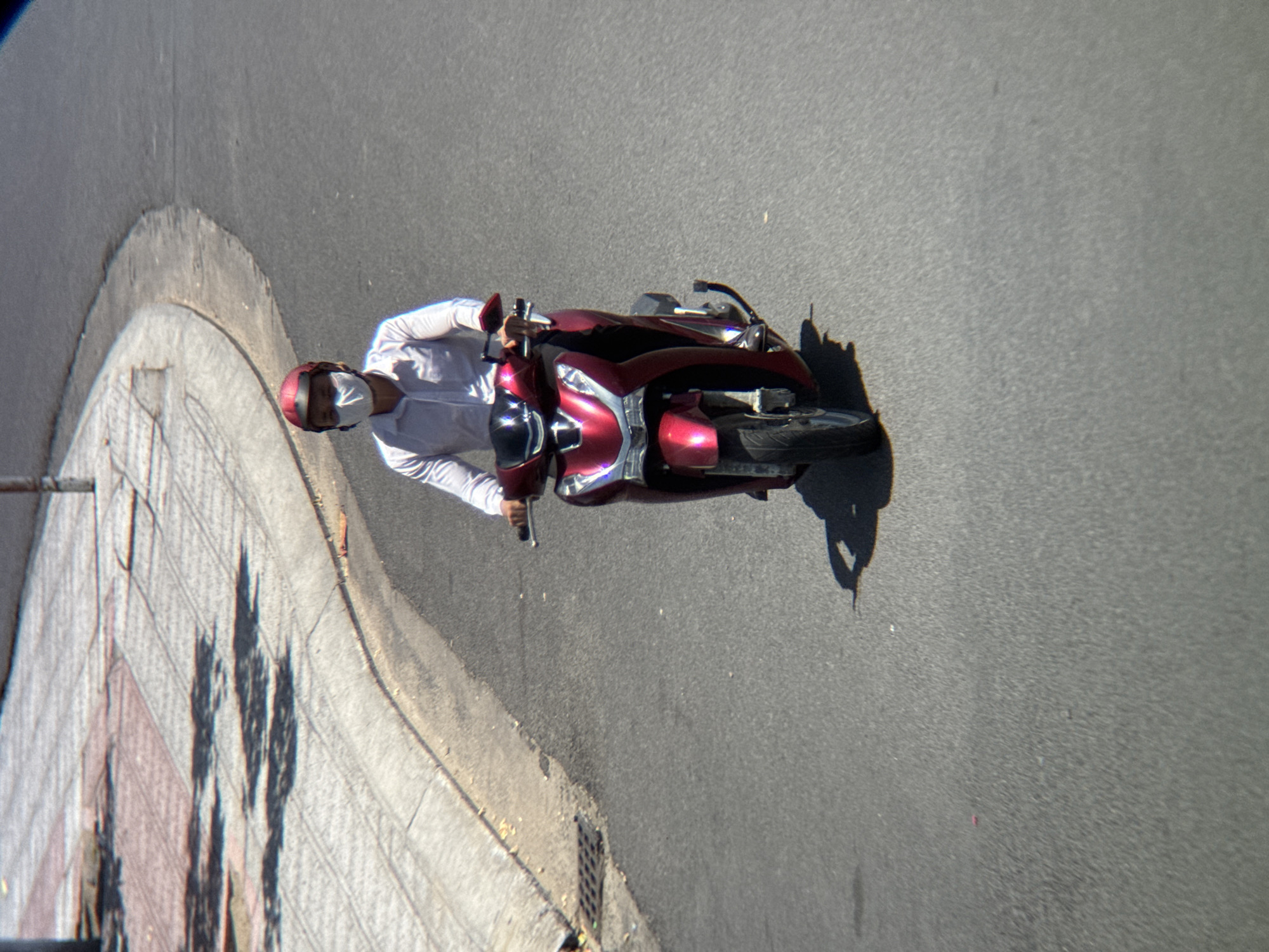 A person wearing a white long-sleeved shirt, a red helmet, and a white face mask is riding a red scooter on a paved road in Nha Trang, Vietnam. The scooter has a silver trim on the front. The road has a curved boundary with a sidewalk on the left, which has a patterned surface and shadows cast from unseen trees. A metal grate is visible on the edge of the sidewalk. The overall lighting suggests it is daytime.
