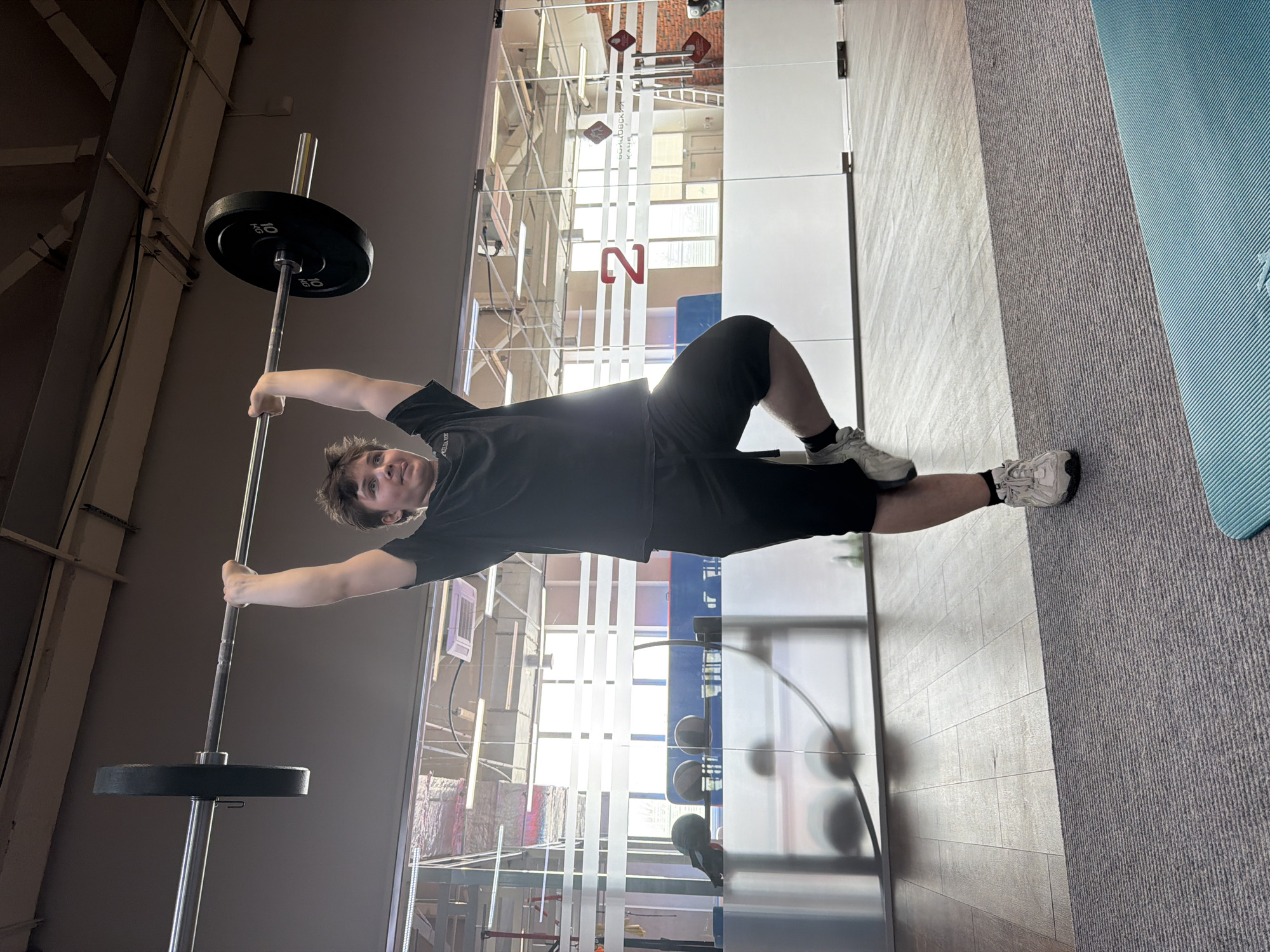 A young man in a black t-shirt and black shorts is holding a barbell with weights in a gym. He is lying on the floor, with his legs bent and feet near a gray and light blue mat. The gym has large glass walls with metal framing. Reflected in the glass are various gym equipment and the interior of a building with visible structural supports and signs in Russian. Based on the location of signs in Russian, the gym is located in Chaikovsky, Russia.