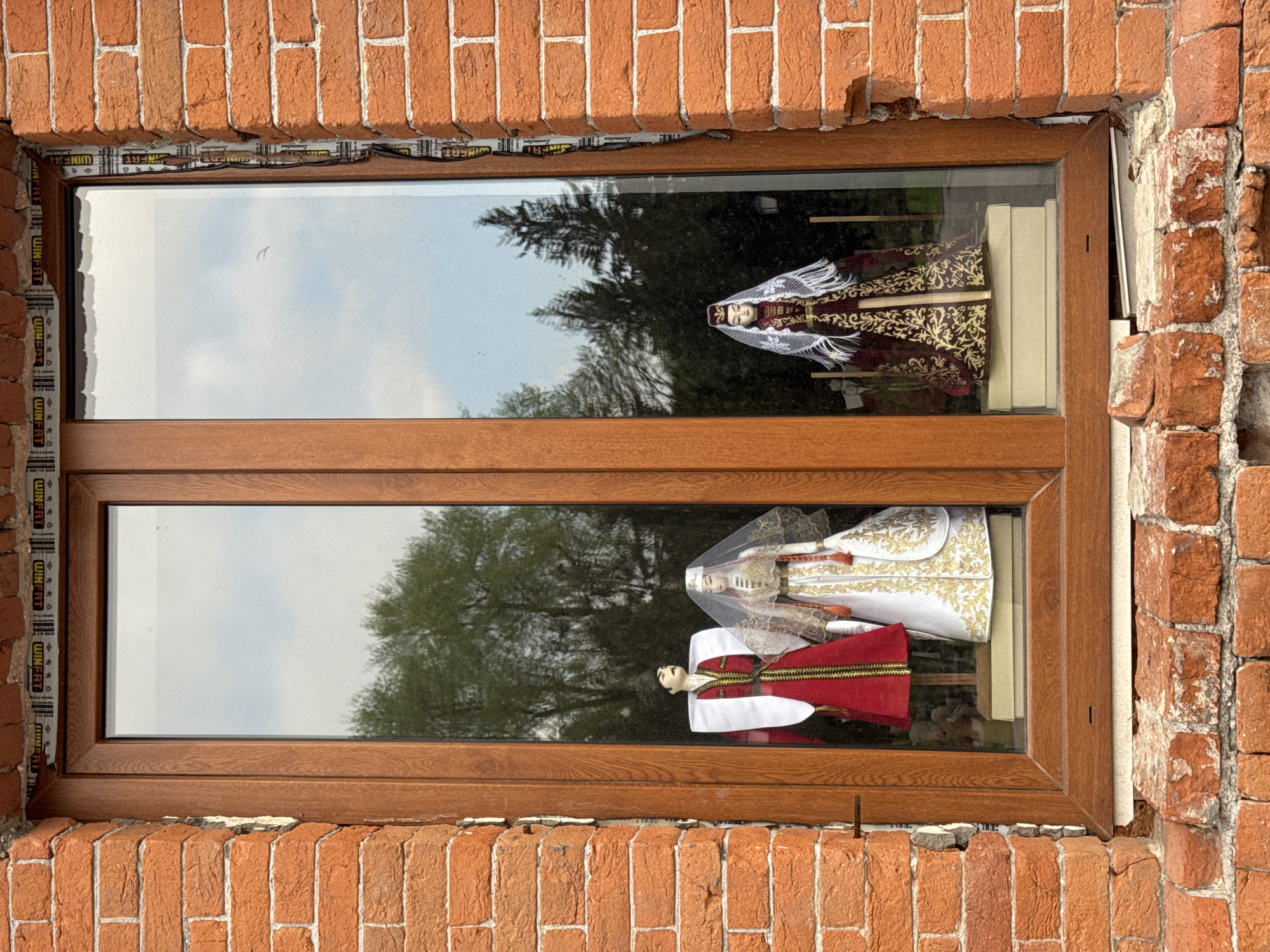 The image shows a window framed by brickwork, likely located in Akhieli, Georgia, given the presence of traditional Georgian dolls displayed inside. The window is a divided pane style with a brown wooden frame. Inside, on a shelf, are three dolls dressed in traditional Georgian attire. The top doll wears a dark, patterned robe with a white lace headdress. The lower dolls include a figure in a white and gold robe with a matching headdress and a figure in a red and white robe. The background visible through the window is a blurred reflection of trees and sky, suggesting an outdoor setting. The brickwork surrounding the window appears aged, adding to the setting’s character.