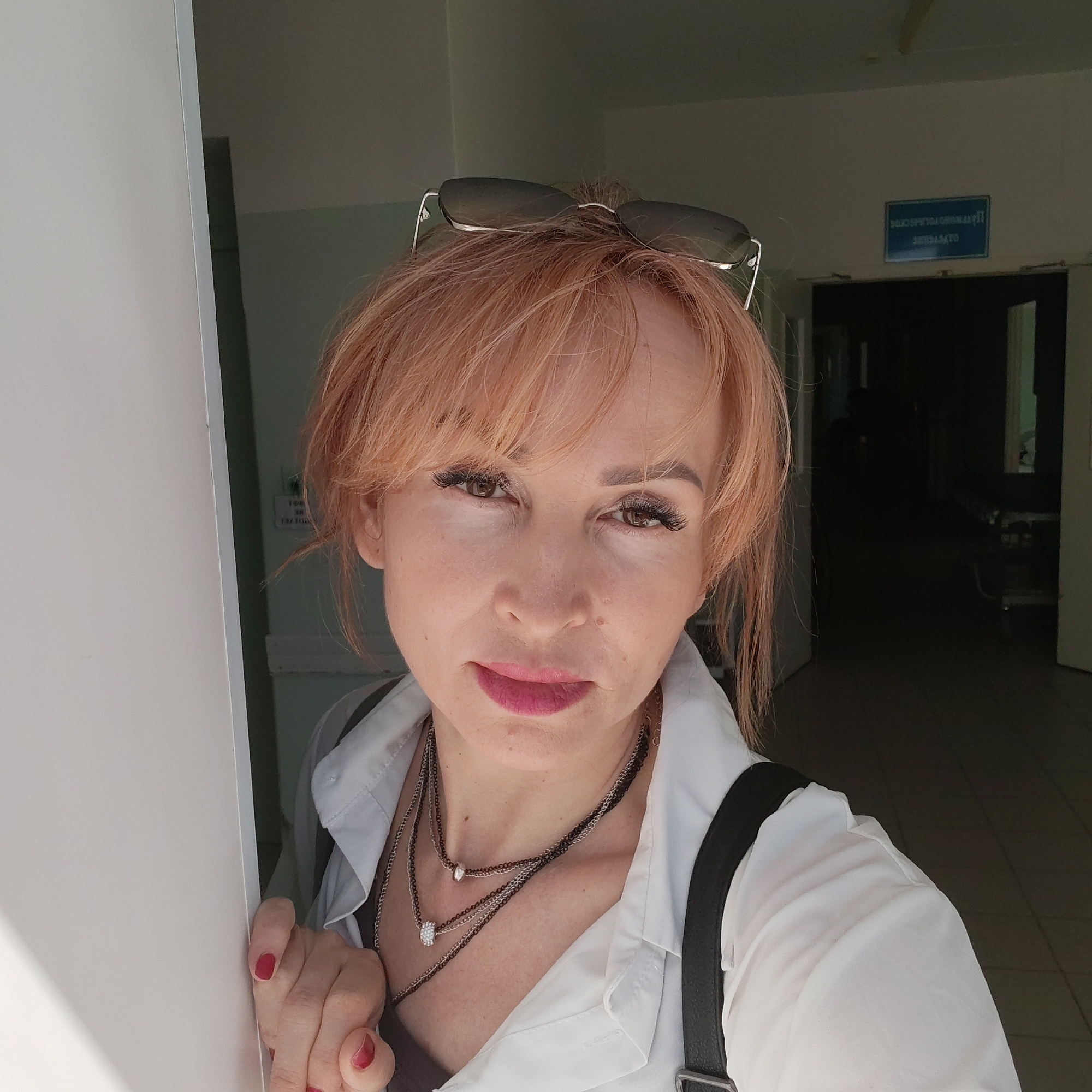 A woman with auburn hair and sunglasses perched on her head stands partially behind a white door, in what appears to be a hallway. She wears a white blouse, a dark-colored shirt underneath, and multiple silver necklaces. A black strap of a bag or purse is visible on her shoulder. Her left hand is visible, holding onto the edge of the white door, and fingernails are painted red. The hallway beyond shows a sign written in Russian Cyrillic letters, indicating a location in the city of Chaikovsky, Russia. The hallway is dimly lit and tiled. There appear to be closed doors and the suggestion of hospital beds or other furniture in the background.