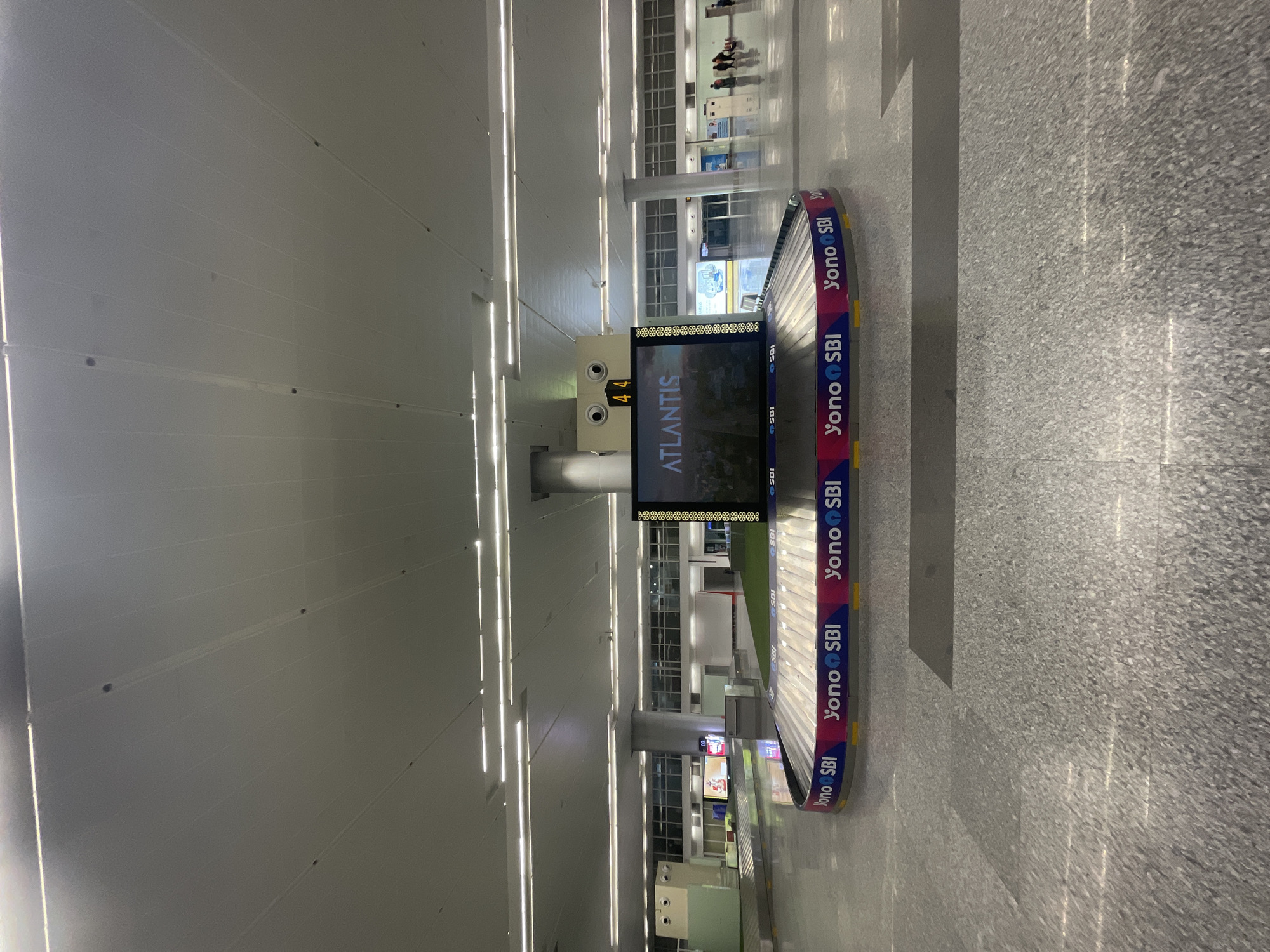 The image depicts an interior view of an airport baggage claim area, specifically at Swami Vivekananda Airport (R new) in Raipur Khurd, India. In the foreground, a baggage carousel is prominently featured, adorned with "Yono SBI" branding. Above the carousel, a large digital display screen shows "ATLANTIS" in large white text, with a smaller number "44" displayed above it. The floor is a polished, speckled gray material. In the background, through large windows, a seemingly empty airport terminal can be seen. A few individuals are visible in the far background, walking. The overall environment is clean, well-lit with fluorescent lighting from the ceiling, and appears to be at a low traffic time.