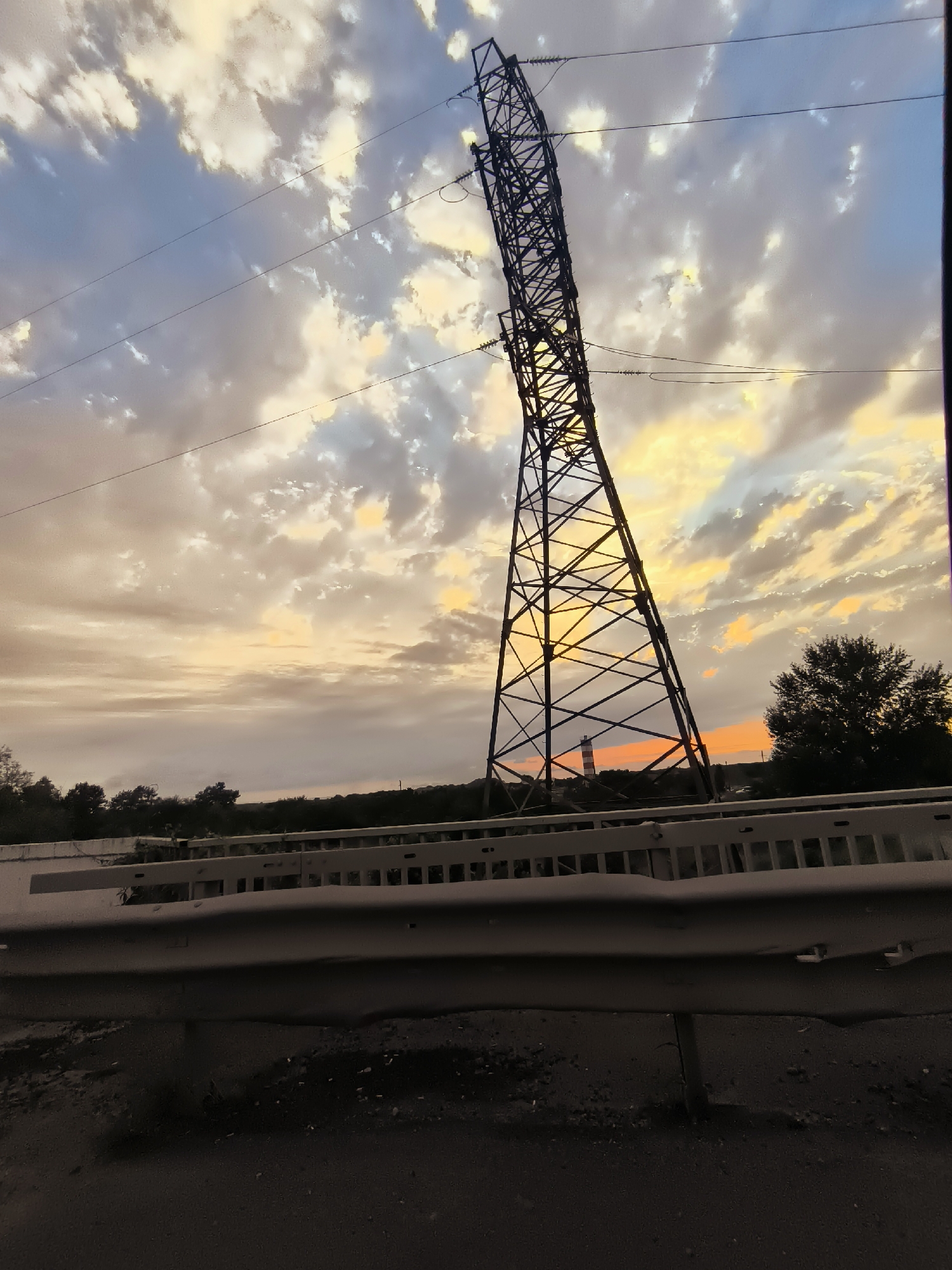 A wide-angle, low-angle shot captures a dramatic sunset in Charnali, Georgia, with a large electricity pylon dominating the right side of the frame. The sky is filled with an array of clouds, ranging from pale yellow and orange near the horizon to shades of blue and grey higher up. Several power lines stretch horizontally across the sky.
In the middle distance, a lighthouse-like structure with red and white bands is visible, nestled amongst dark trees. The foreground is occupied by a light-colored metal guardrail, likely from a bridge or overpass, with dark asphalt or road surface below. The overall scene suggests a rural or semi-rural setting at dusk.