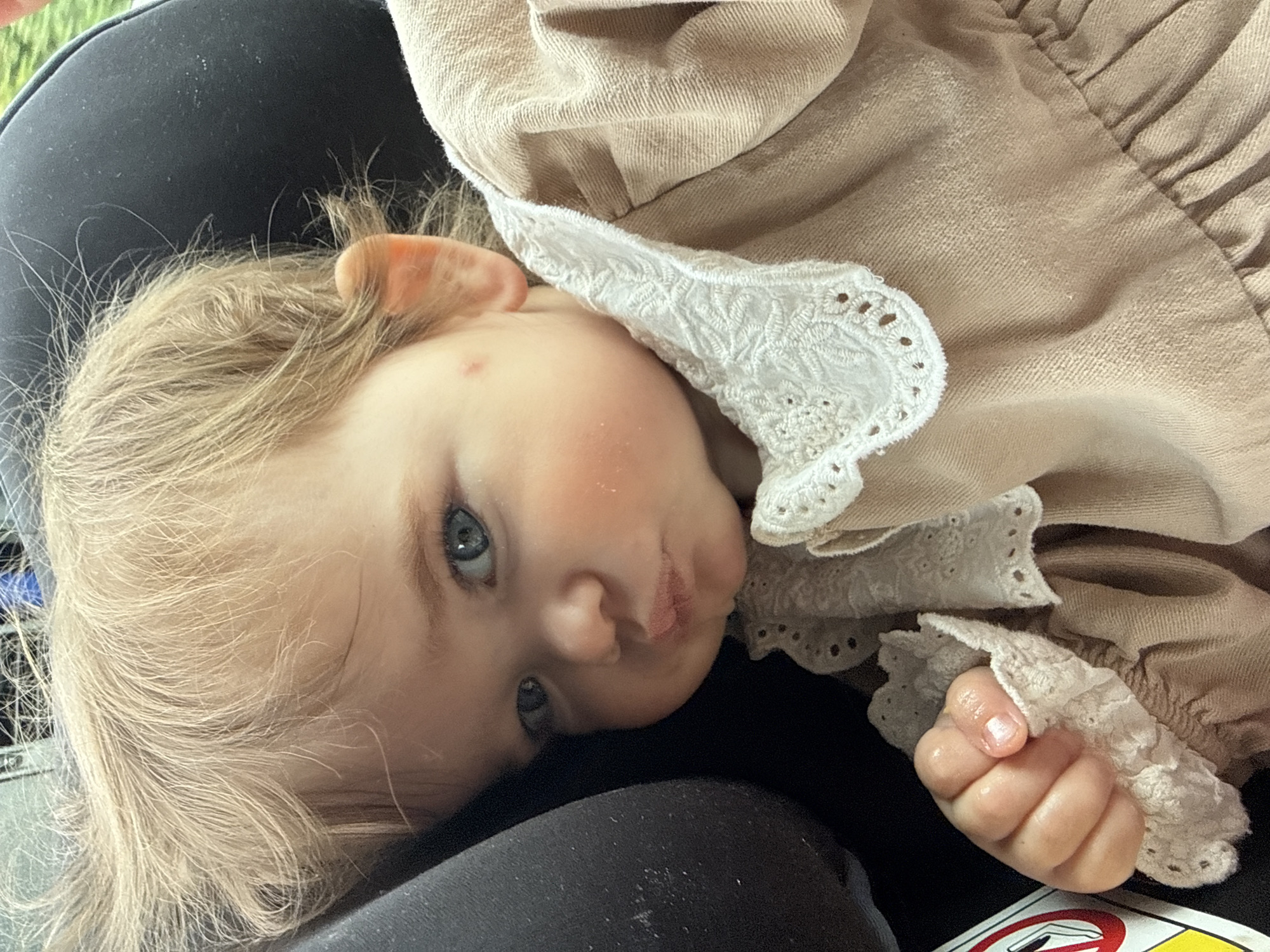 A close-up, eye-level shot shows a baby with blond hair and blue eyes. The baby is wearing a tan dress with white lace trim. The baby's left hand is visible, holding onto the lace. The baby is in a car seat, indicated by the black headrest and a portion of a warning label visible on the left. The lighting suggests it is daytime, possibly outdoors. The image is taken in Limburg an der Lahn, Germany.