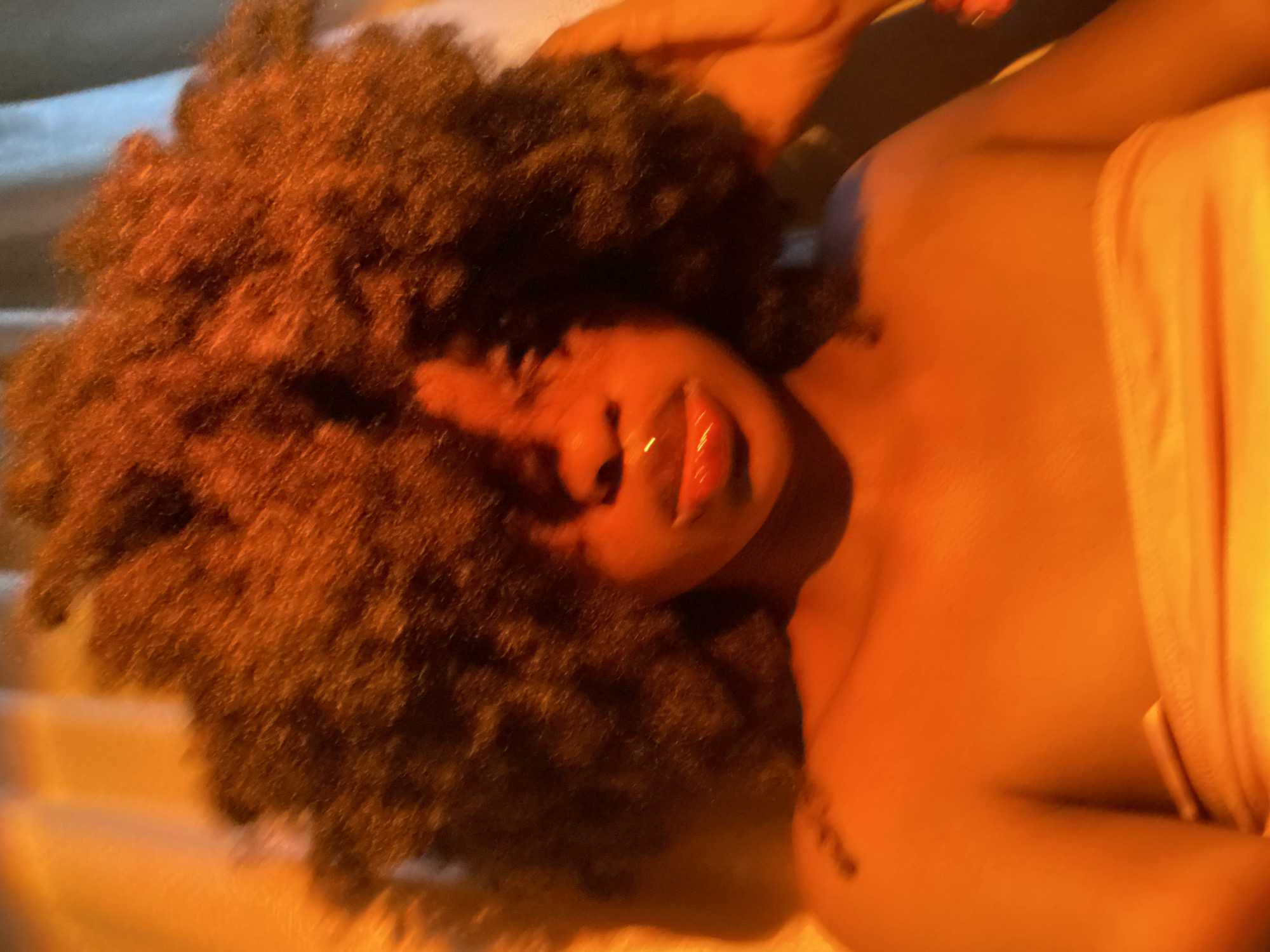 A low-angle studio shot depicts a brown-skinned woman with dark brown, voluminous afro-textured hair. She is reclined, seemingly on a surface with linear indentations. Her lips appear glossy. Her body is visible from the shoulders up, with fabric draped around her. The lighting creates an overall golden-orange hue. There is no indication of Iranla, Nigeria.