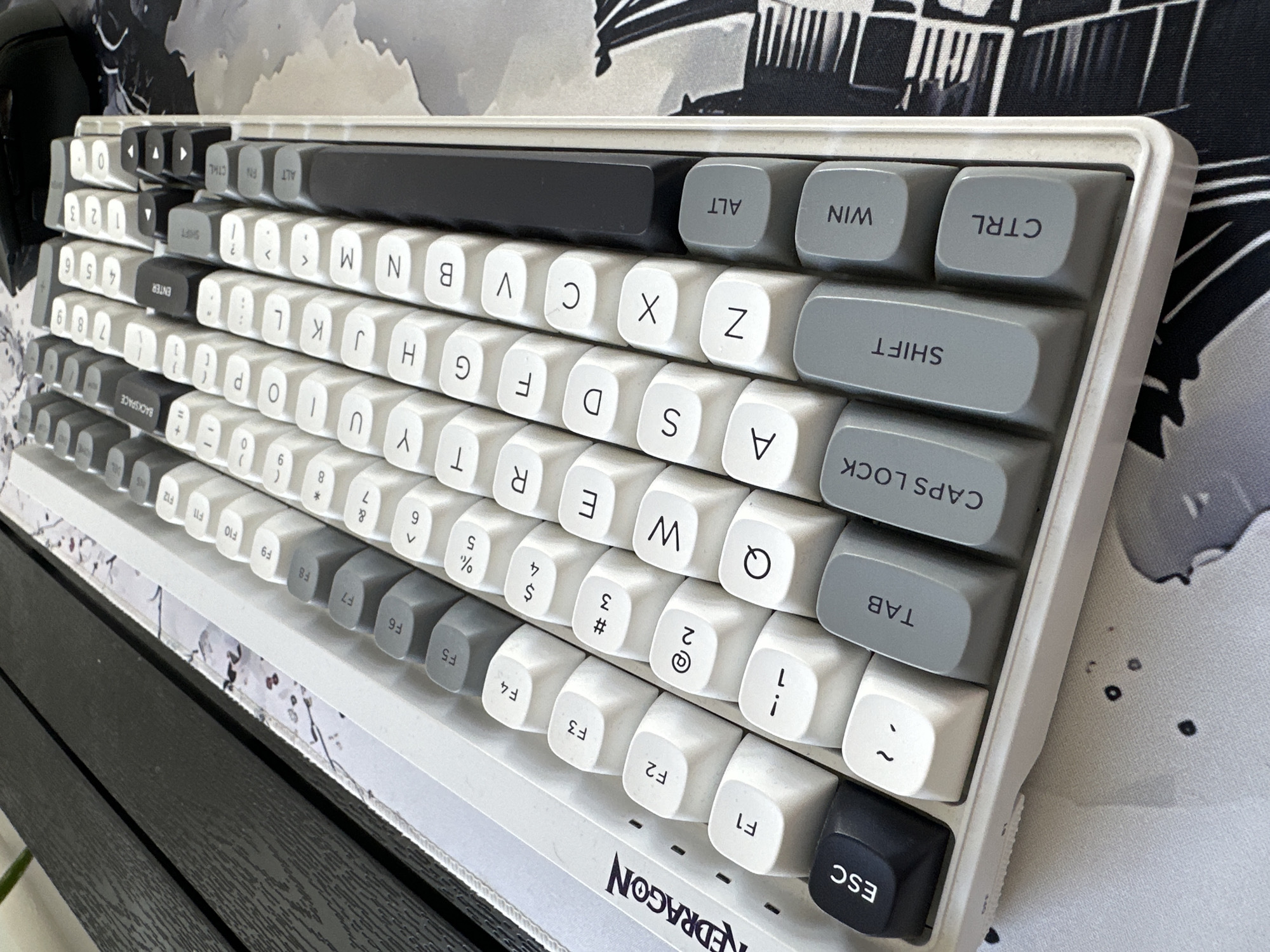 The image shows a close-up of a white mechanical keyboard with a two-tone keycap design, featuring white and grey keys. The keyboard is positioned on a desk with a dark wooden surface to the left and a patterned desk mat with grey and white abstract designs underneath the right side. The Redragon logo is visible on the top left edge of the keyboard. The keyboard is angled, with the top right corner closer to the viewer. Function keys (F1-F12) are arranged in the top row, followed by number keys, and then the alphanumeric keys. Modifier keys such as ESC, TAB, CAPS LOCK, SHIFT, CTRL, WIN, and ALT are visible on the left and bottom rows. The keys are illuminated by ambient light, casting subtle shadows. No people are present in the image. The setting appears to be an indoor workspace. The specific location of Moortown, Ireland, is not evident from the image itself.