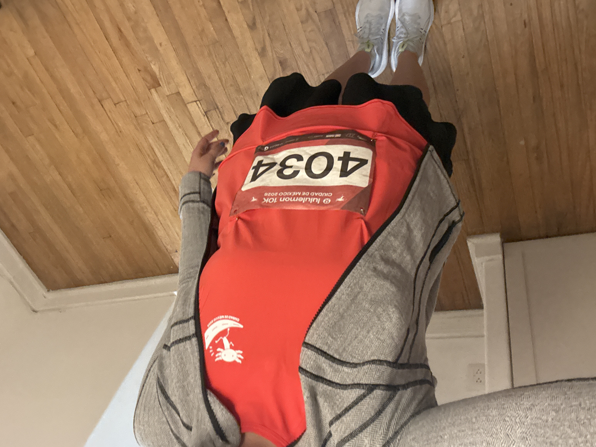 The image shows a person wearing a red shirt with a white logo featuring an axolotl under a mushroom. The shirt also has a race number "4034" and the words "lululemon 10K CIUDAD DE MEXICO 2026". The person is wearing a gray jacket. The picture seems to have been taken indoors; a wooden floor is visible. The information on the shirt indicates the location is Mexico City, Mexico.