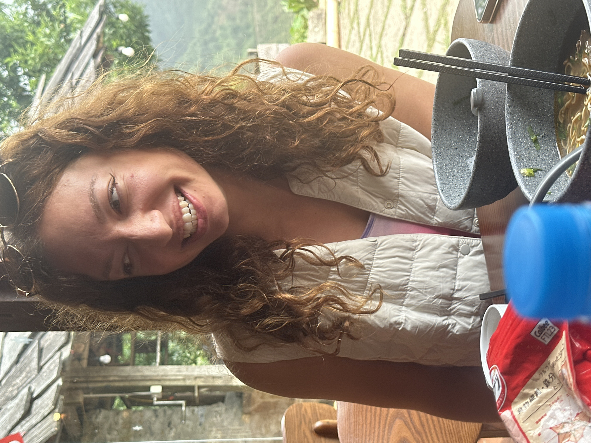 A young Caucasian woman with curly brown hair is smiling widely while looking to her left. She is wearing a light-colored sleeveless puffy vest over a pink and purple top. Her sunglasses are perched on top of her head. The background appears to be an outdoor seating area with a wooden table and chairs, suggesting a cafe or restaurant. In the foreground, there are two bowls of ramen with chopsticks and a red package of what looks like snacks, and a blue bottle cap. The setting suggests a rural mountainous region, consistent with Ta Xua, Vietnam, known for its misty landscapes and ethnic minority villages.