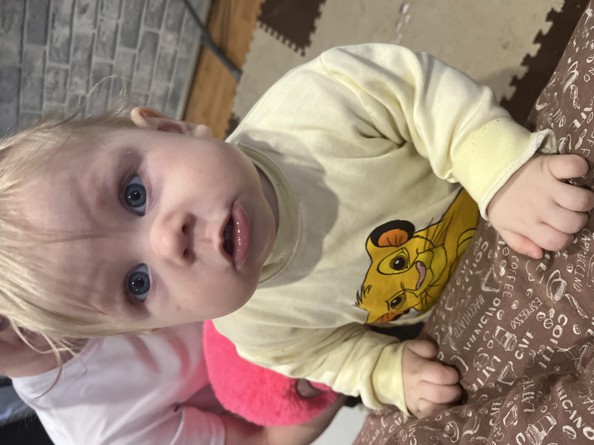 A young child, likely a baby or toddler, is captured in a close-up, high-angle shot within an indoor setting. The child has light blonde hair, striking blue eyes, and fair skin, looking directly at the camera with a curious expression and slightly open mouth. They are wearing a pale yellow, long-sleeved shirt featuring a large, colorful print of Simba, the cartoon lion character from Disney's The Lion King.
In the background, a grey wall with a rectangular brick-like pattern is visible, alongside a floor covered with interlocking brown foam mats, suggesting a play area. Part of an adult's white shirt is seen in the lower-left corner, indicating the child is near or being held by an adult. The child's right arm rests on a brown fabric, possibly a blanket or cushion, which is adorned with white illustrations of coffee cups and various coffee-related terms. Visible text on this fabric includes "CAPPUCCINO," "ESPRESSO," "COFFEE," "LATTE," "MACCHIATO," "AMERICANO," and "CAFE." A bright pink, fluffy object is partially visible beneath the child's left side. The scene is brightly lit, focusing intently on the child.