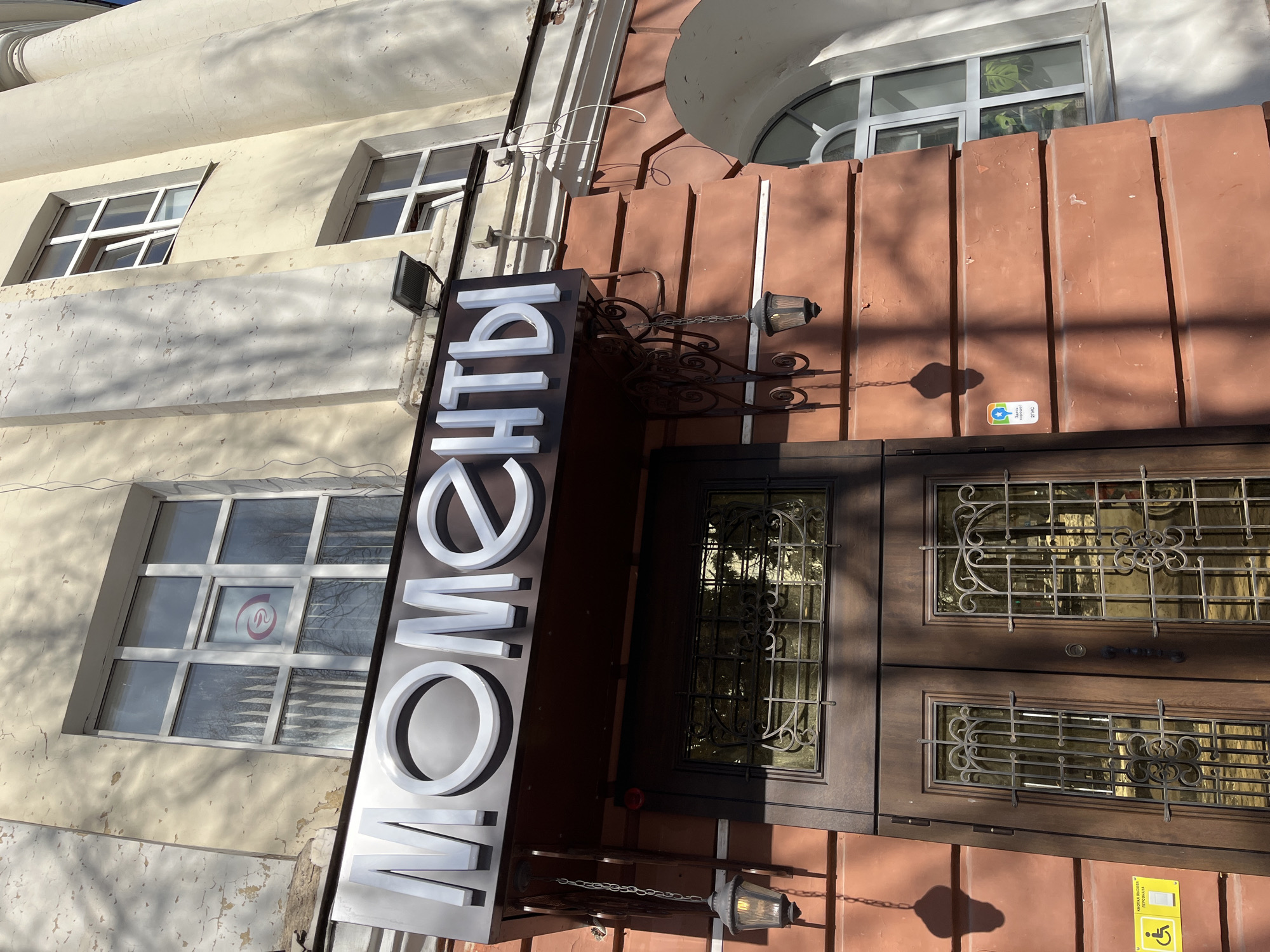 The image shows the entrance to "Momenti" in Izhevsk, Russia. A sign with the word "Моменты" (Momenti) is displayed vertically on the left of the entrance, with white letters. The building exterior has two different colored sections: the left side is light yellow, while the entrance area on the right is a reddish-brown. There are two windows visible on the yellow side and one arched window on the reddish-brown side. The double door entrance is brown, with decorative metal grillwork covering windows in the doors. A wrought-iron bracket holding a vintage-style lantern hangs above the door. Two circular plaques are attached to the reddish-brown wall, with the lower plaque containing an icon of accessibility.