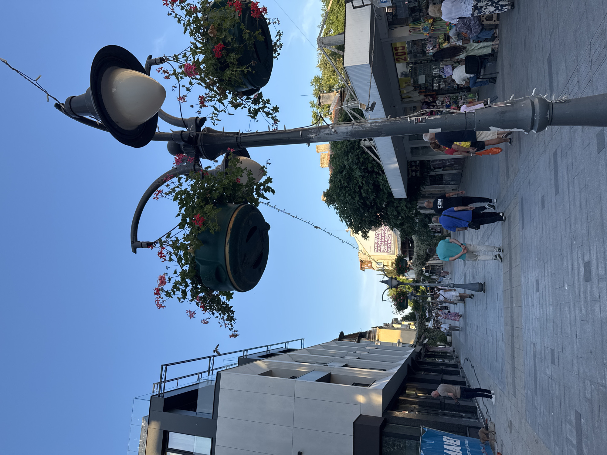The image depicts a wide pedestrian street in Burgas, Bulgaria, likely during daytime based on the clear blue sky and natural lighting. The street is paved with grey interlocking pavers and lined with shops and buildings. On the left, a modern building with light-colored facade and dark accents, featuring balconies and large windows, stands prominently. Further down the street on the right, a row of shops with awnings and displays of merchandise is visible, including one with a sign indicating prices like "200".
Several people are walking along the street. Towards the left, a man in a light-colored shirt and dark pants is standing next to a large blue banner that reads "MAVE". A small dog is near him. Further down the street, a group of people are strolling, some are couples, others are families. A man in a green shirt and khaki pants is walking away from the camera, as is a man in a black t-shirt with "CSM" printed on it. People of various ages and attire are present, some are carrying bags.
Decorative elements include street lamps with hanging flower baskets filled with red flowers, and string lights draped across the street. Trees and foliage are visible in the background, suggesting a pleasant urban environment. The overall scene conveys a lively atmosphere of a shopping and pedestrian zone.