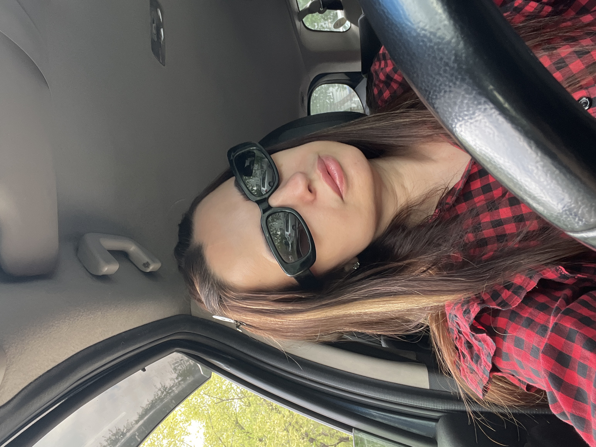 A woman is in the driver's seat of a car. She is wearing sunglasses and a red and black checkered shirt. The car is in an unknown location. The driver's side window is down. Outside, green trees are visible.