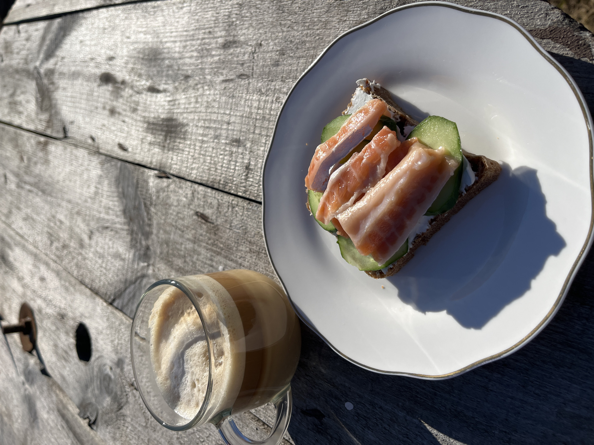 The image shows a wooden picnic table outdoors, with a white plate containing an open-faced sandwich. The sandwich is comprised of a dark bread base, covered with white cream cheese, slices of cucumber, and topped with slices of salmon. Next to the plate is a clear glass mug filled with a foamy, light-brown beverage that appears to be a latte or cappuccino. The setting is a sunny outdoor location in the city of Chaikovsky, Russia.