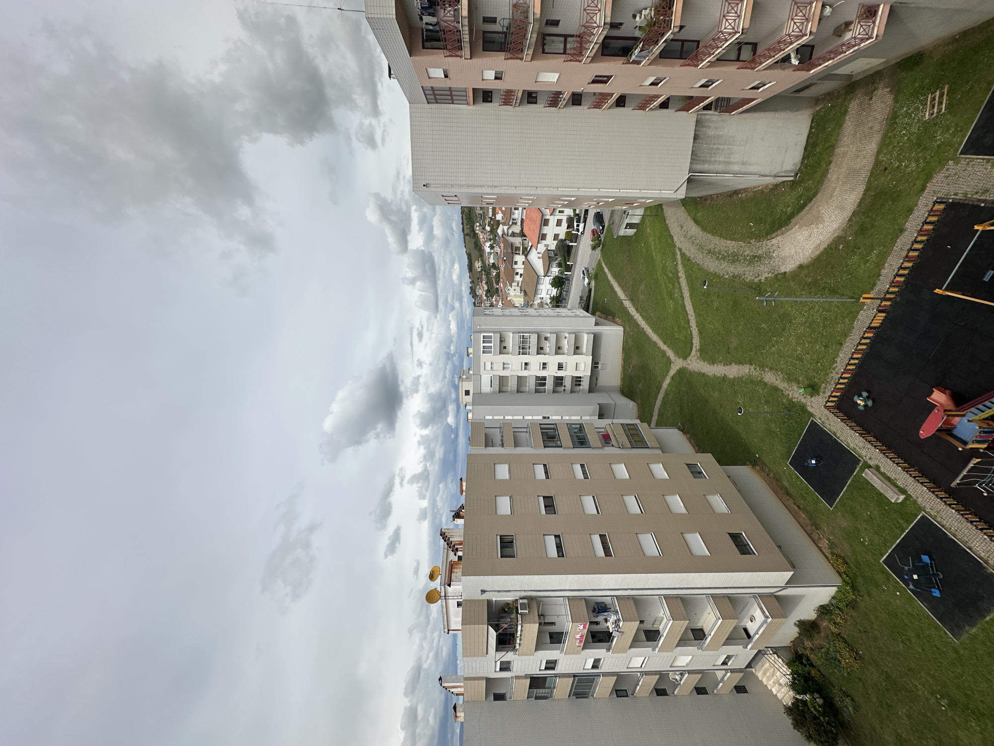 This image shows a view of residential buildings in Bragança, Portugal, likely taken from a high vantage point such as an upper floor window. The sky is overcast with grey and white clouds. The buildings are varying heights and colors, including beige, white, and light pink, and feature balconies and rectangular windows. A playground with black rubber surfaces, climbing equipment, and other play structures is visible in the lower right, surrounded by green grass. Paths, made from light colored small stones or gravel, run through the grassy areas between the buildings. The horizon reveals houses and trees in the background. There are no people visible in the shot. Satellite dishes can be seen mounted on one of the buildings.