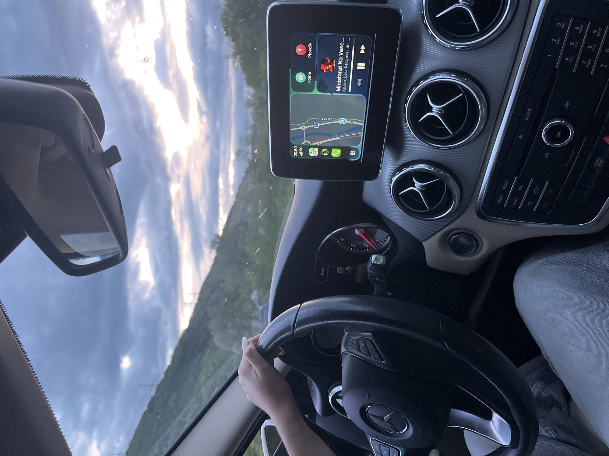 The image shows the interior of a Mercedes-Benz car, with the driver's hand on the steering wheel. The dashboard features a screen displaying Apple CarPlay, showing a map indicating the location near Struma river, close to the city of Blagoevgrad, Bulgaria. The CarPlay interface also shows that the driver is currently listening to the song "Ministarat Na Veselieto" by Krisko, Lora Karadjova and Boris Dali. The time displayed on the car's screen is 20:50, and the phone is connected to 5G. The image also captures the car's air vents, central console, and a portion of the rearview mirror. Through the windshield, there's a view of the sky with scattered clouds and the outline of a landscape, possibly trees or hills. The overall lighting suggests it's either late afternoon or early evening.
