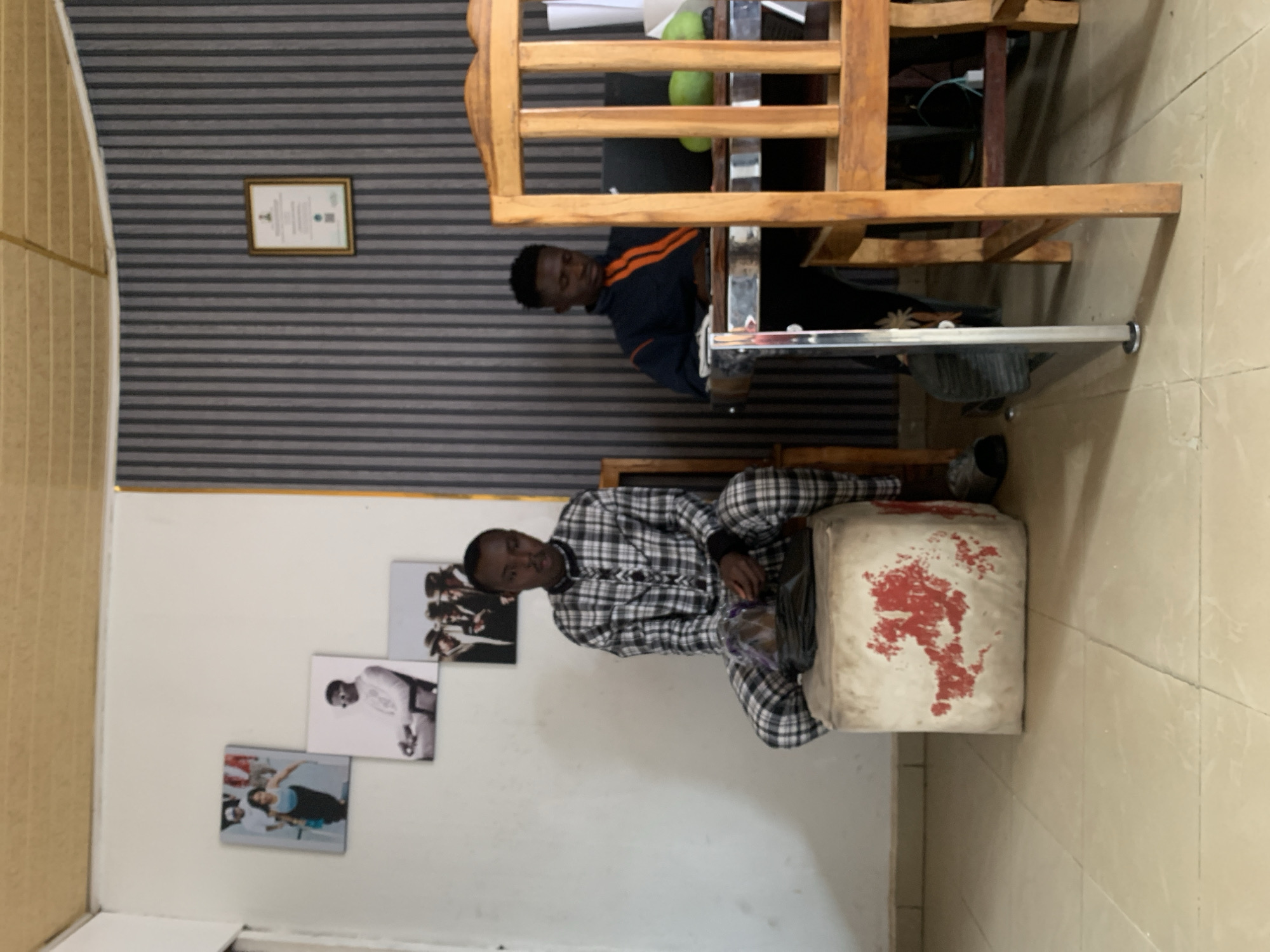 The image depicts an indoor scene, likely in an office or study, possibly located in Jos, Nigeria. Two young men are present. One man, wearing a black and white checkered outfit, is sitting on a white cube-shaped object with red markings. The second man, wearing a dark jacket with orange stripes, is seated at a wooden desk, partially obscured by its structure. Behind them, the wall is decorated with three framed pictures and a framed certificate hanging on the wall with grey and black horizontal stripes. The floor is tiled with a light tan material.