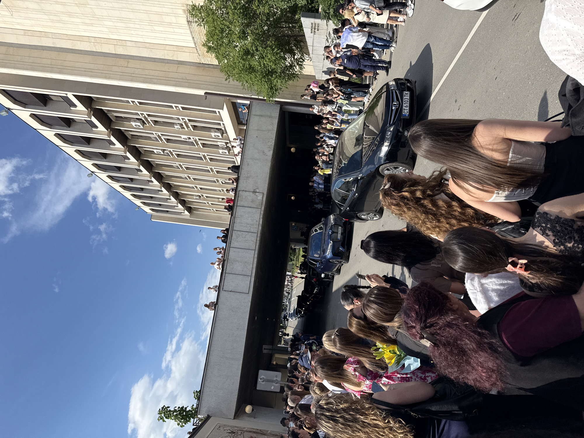 The photograph shows a crowd of people gathered along a street with a bridge or overpass. Cars are passing through the underpass. The crowd is mostly composed of young women, some of whom are holding flowers. Many people are observing and using their phones to take pictures or videos. A multi-story building is on the left side of the frame, with people visible at some of the windows. The sky is blue with scattered clouds. There is a license plate visible on a black car of CT 6103 TK. Based on the style of license plate and architectural features, the location is Stara Zagora, Bulgaria.