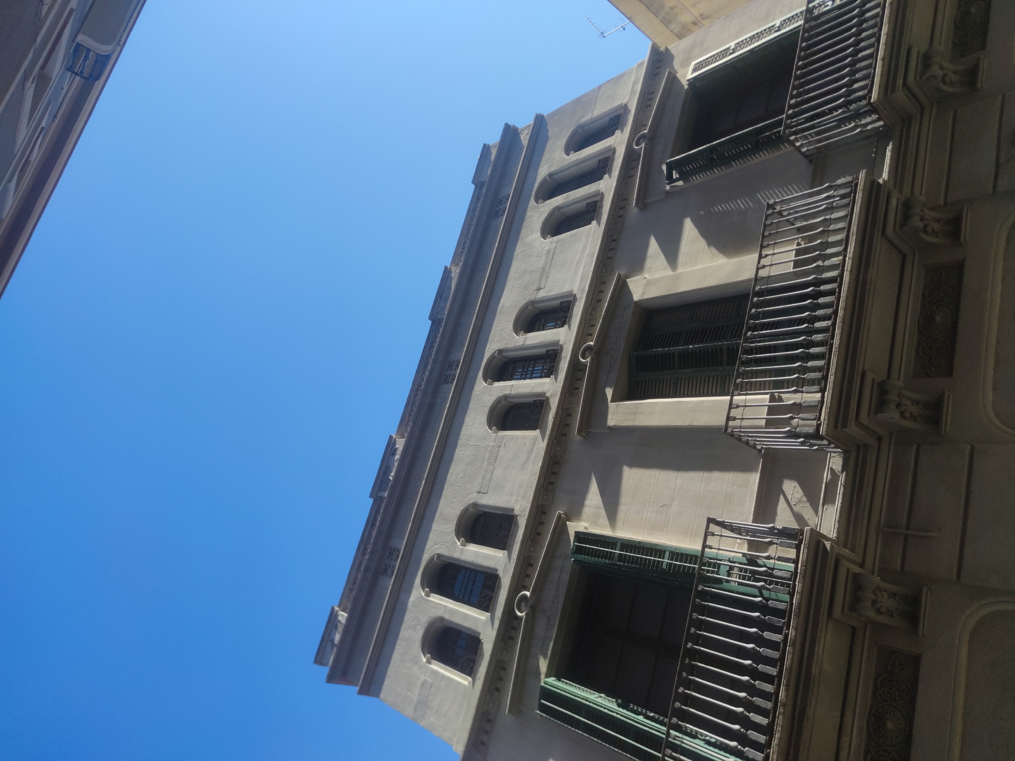The image shows a tall, light-colored building with multiple stories, viewed from a low angle. The building has a symmetrical design with arched windows on one side and windows with grated balconies on the other. The sky is a clear, bright blue. The city location is unknown.