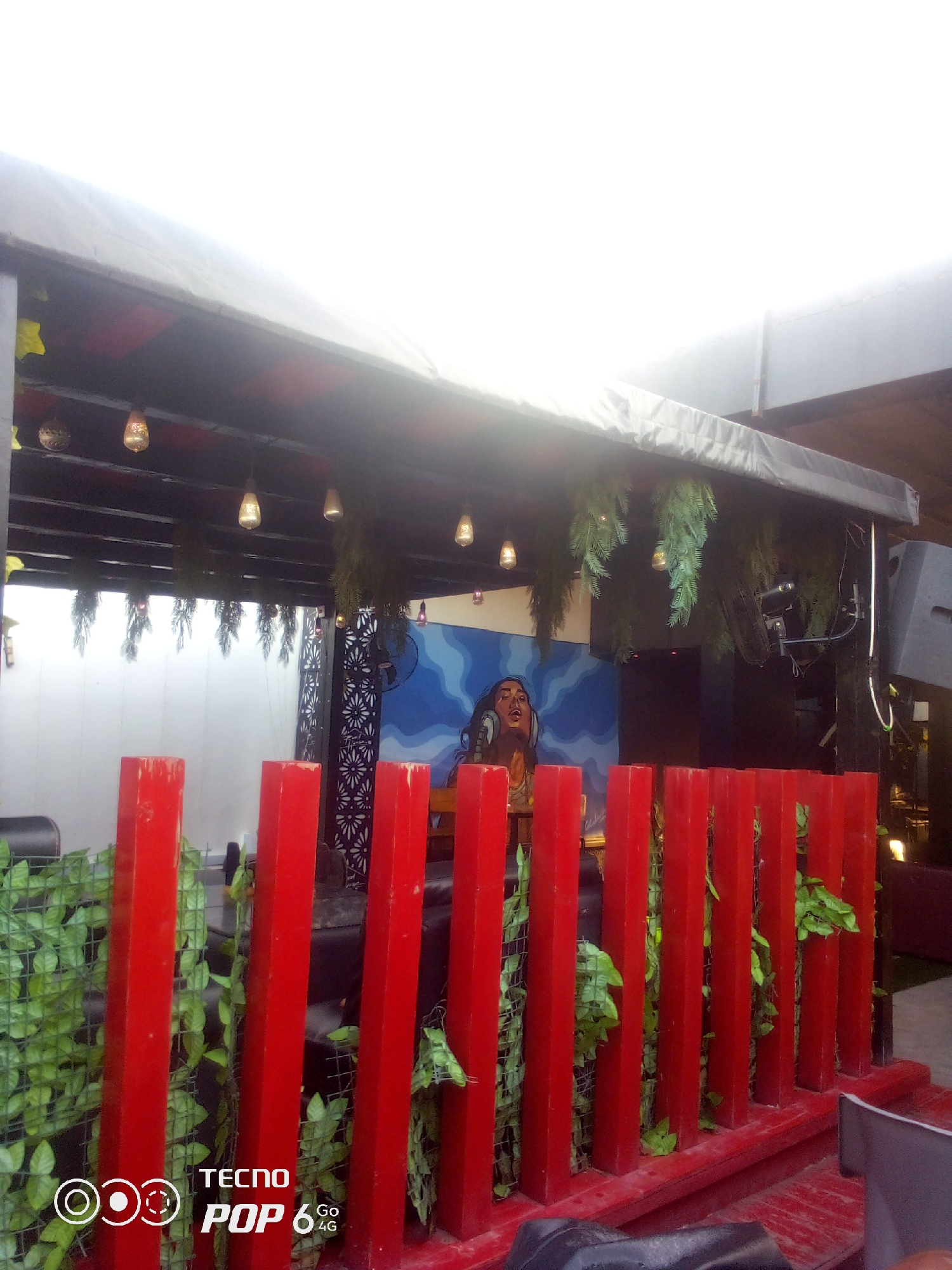 The image shows an outdoor entertainment venue, possibly a bar or lounge, in Howayi, Nigeria. A red wooden fence adorned with artificial green foliage runs across the foreground. Behind the fence, a DJ booth is visible, featuring a mural of a person with headphones against a blue, wavy background. Several hanging lights illuminate the covered ceiling of the venue, and tropical plants are suspended from it. Speakers are mounted on a wooden post to the right, and a large black speaker is positioned further back. The overall atmosphere suggests a vibrant and modern setting for nightlife or events.