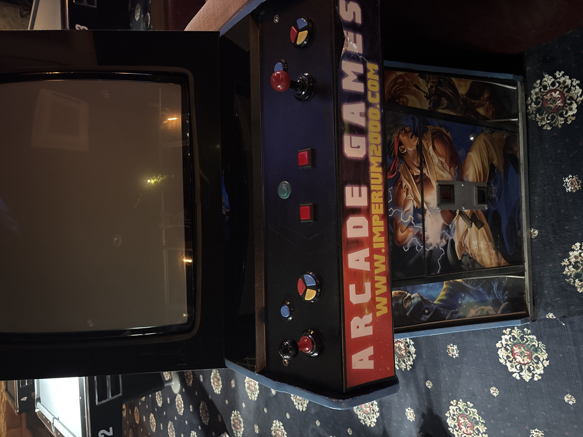 The image shows an "Arcade Games" cabinet, potentially for a retro video game, with a visible screen and control panel featuring buttons and a joystick. The cabinet is decorated with artwork depicting characters in action, along with the URL "www.imperium2000.com." The cabinet is set against a patterned carpet. There is no information to imply the location is in Plovdiv, Bulgaria. No people are visible in the image.