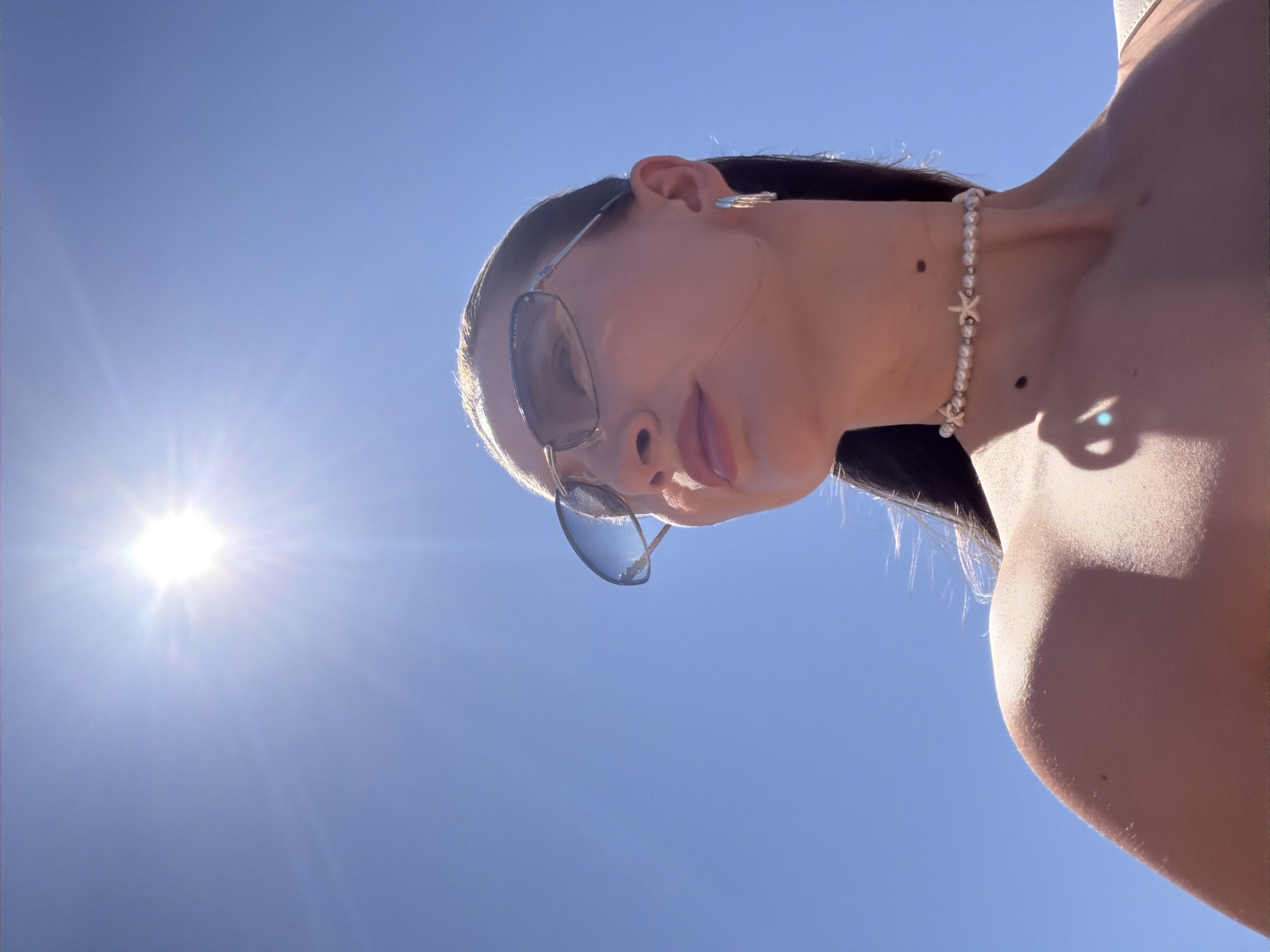 A low-angle shot captures a woman wearing sunglasses and a pearl necklace with starfish charms, silhouetted against a bright blue sky and the sun. The sun is positioned in the upper left quadrant of the image, emitting strong rays that create a lens flare. The woman's face is turned to the right, with her lips slightly parted. She has dark hair pulled back, and is wearing a light-colored top. The overall scene appears to be outdoors, likely on a sunny day.