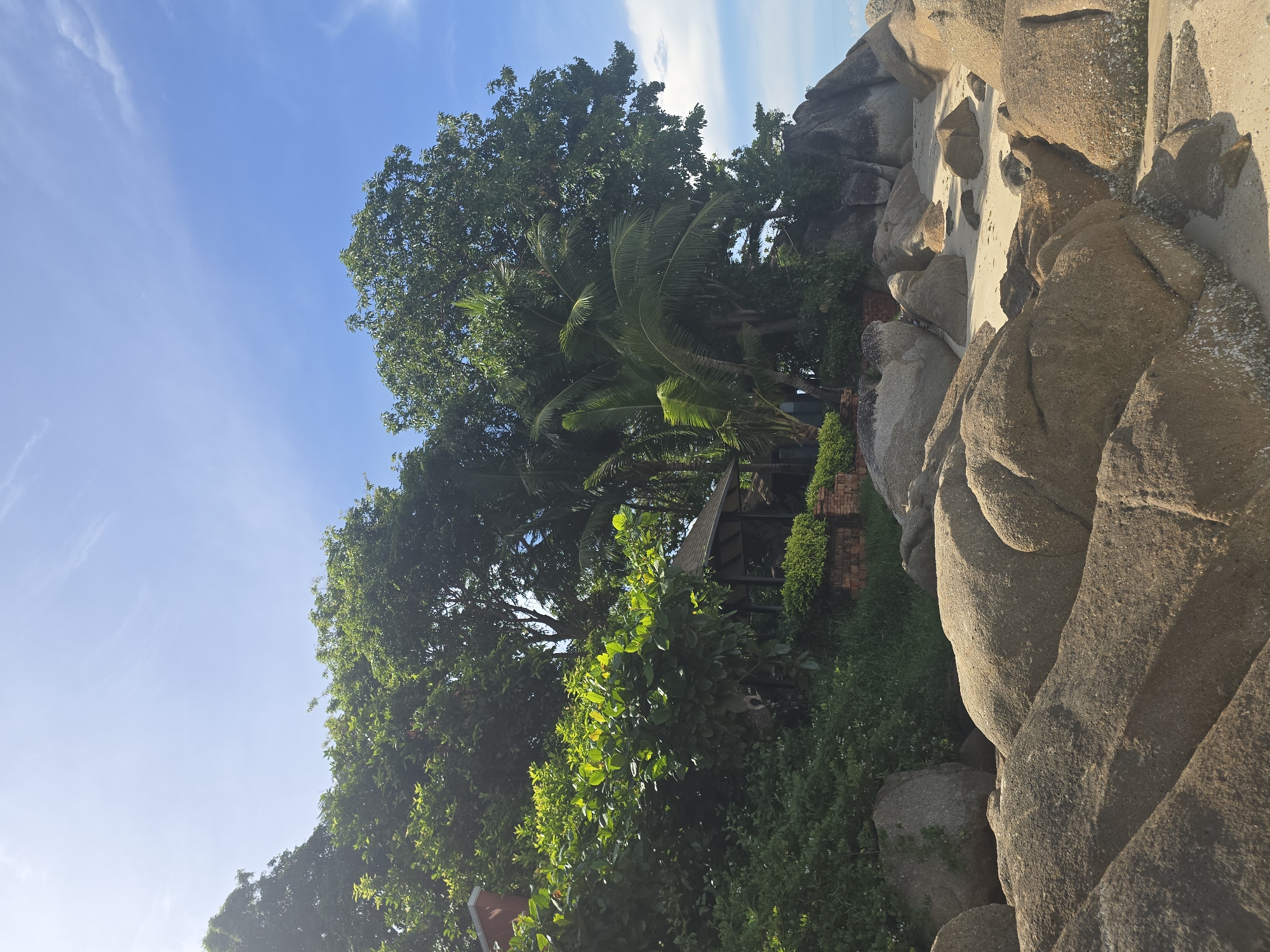 The image depicts a tropical beach scene with large, smooth boulders in the foreground, leading to a sandy stretch of beach. Behind the boulders, lush green vegetation, including trees and palm fronds, dominates the midground. A small, open-sided structure with a dark roof is partially visible amidst the foliage, possibly a gazebo or shelter. Some red brickwork is also visible at the base of the vegetation. The background features a clear blue sky with sparse white clouds. The location is identified as Baan Chaweng Noi, Thailand. There are no people visible in the image.
