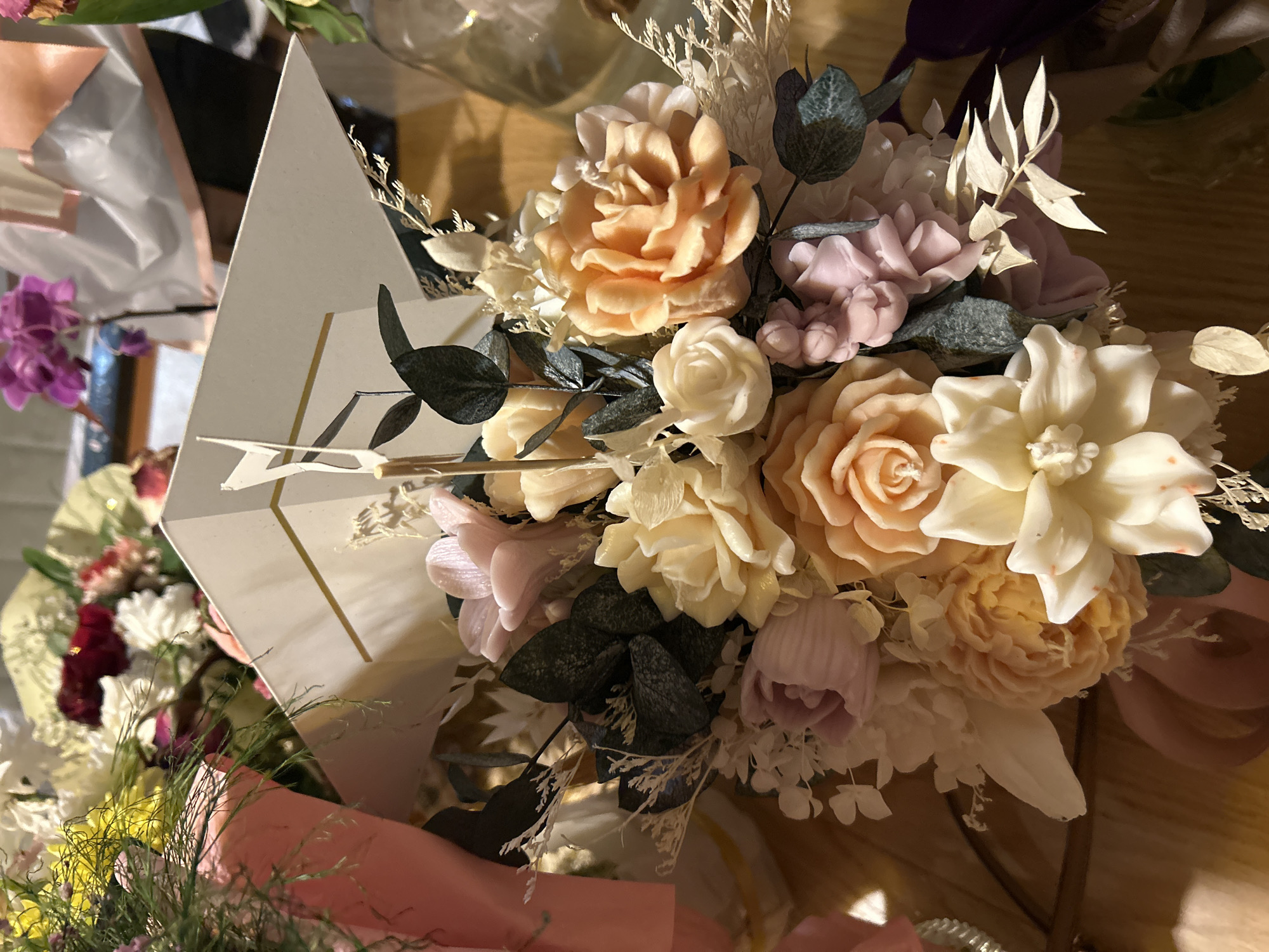 The image shows a bouquet of artificial flowers in pastel shades. The flowers are primarily roses and other similar blooms in peach, cream, white, and light purple. Dark green leaves are interspersed amongst the flowers, and dried floral elements add texture. The bouquet is wrapped in white paper with gold stripes, suggesting a modern and minimalist design. The photo appears to be taken indoors, with a wooden surface visible in the background. Other flowers and objects are partially visible in the periphery, indicating the setting might be a floral shop or a home. There is no evidence that indicates this photo was taken in Burgas, Bulgaria.