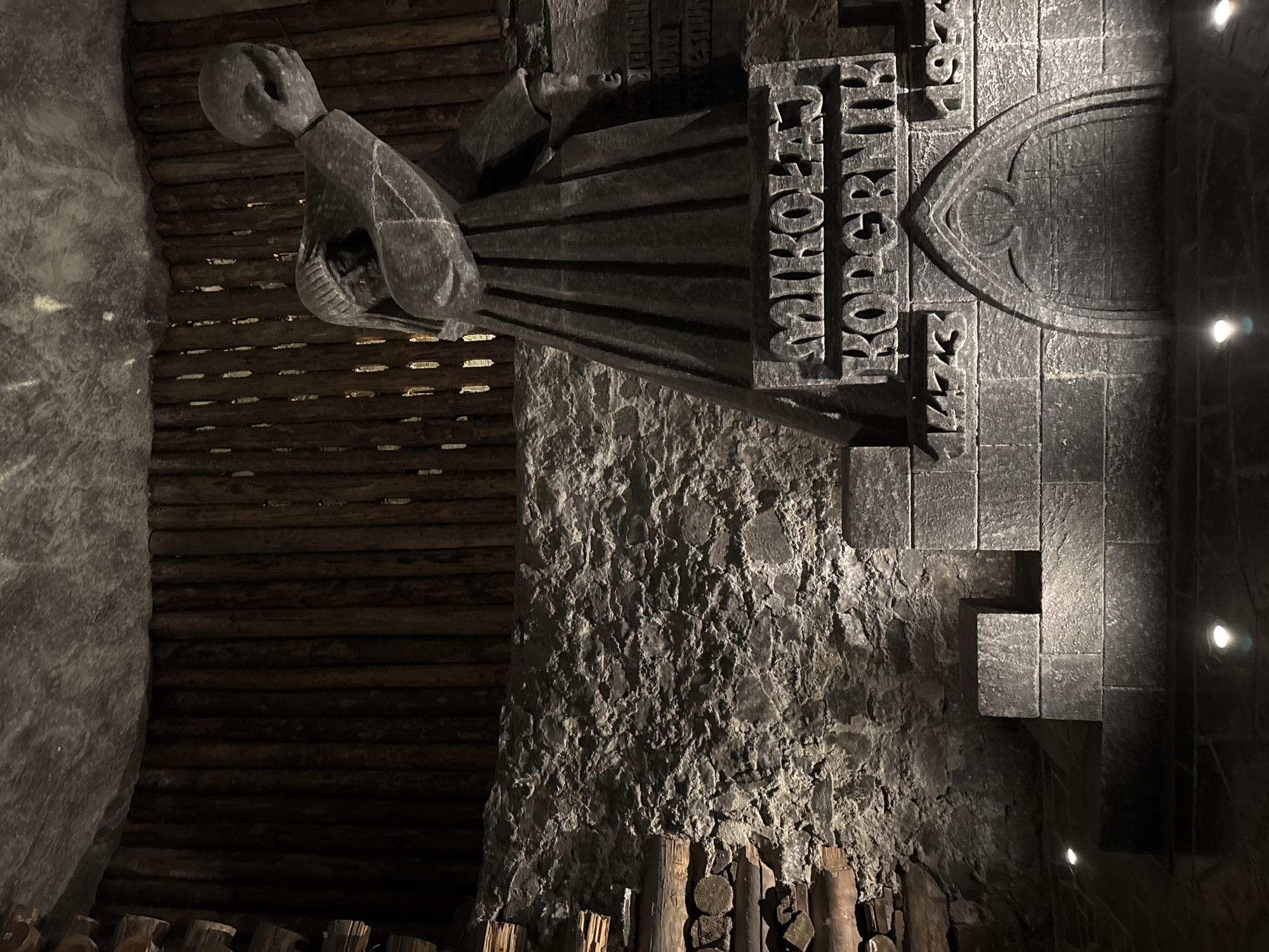 The image shows a statue of Nicolaus Copernicus inside the Wieliczka Salt Mine in Wieliczka, Poland. The statue is carved from salt and depicts Copernicus holding an astronomical instrument. Behind the statue is a wall also carved from salt, with an inscription that includes his name. The statue is set in a chamber lined with wooden logs and stone, illuminated by spotlights.