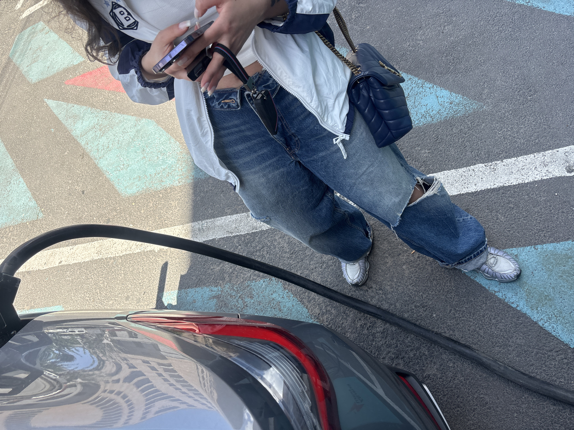 A person in blue jeans and a white jacket is standing next to a gray car that is plugged into a charging station. The person is holding a phone in their hands. The scene appears to be in a parking lot with painted parking spaces. The city and country are Yerevan, Armenia.