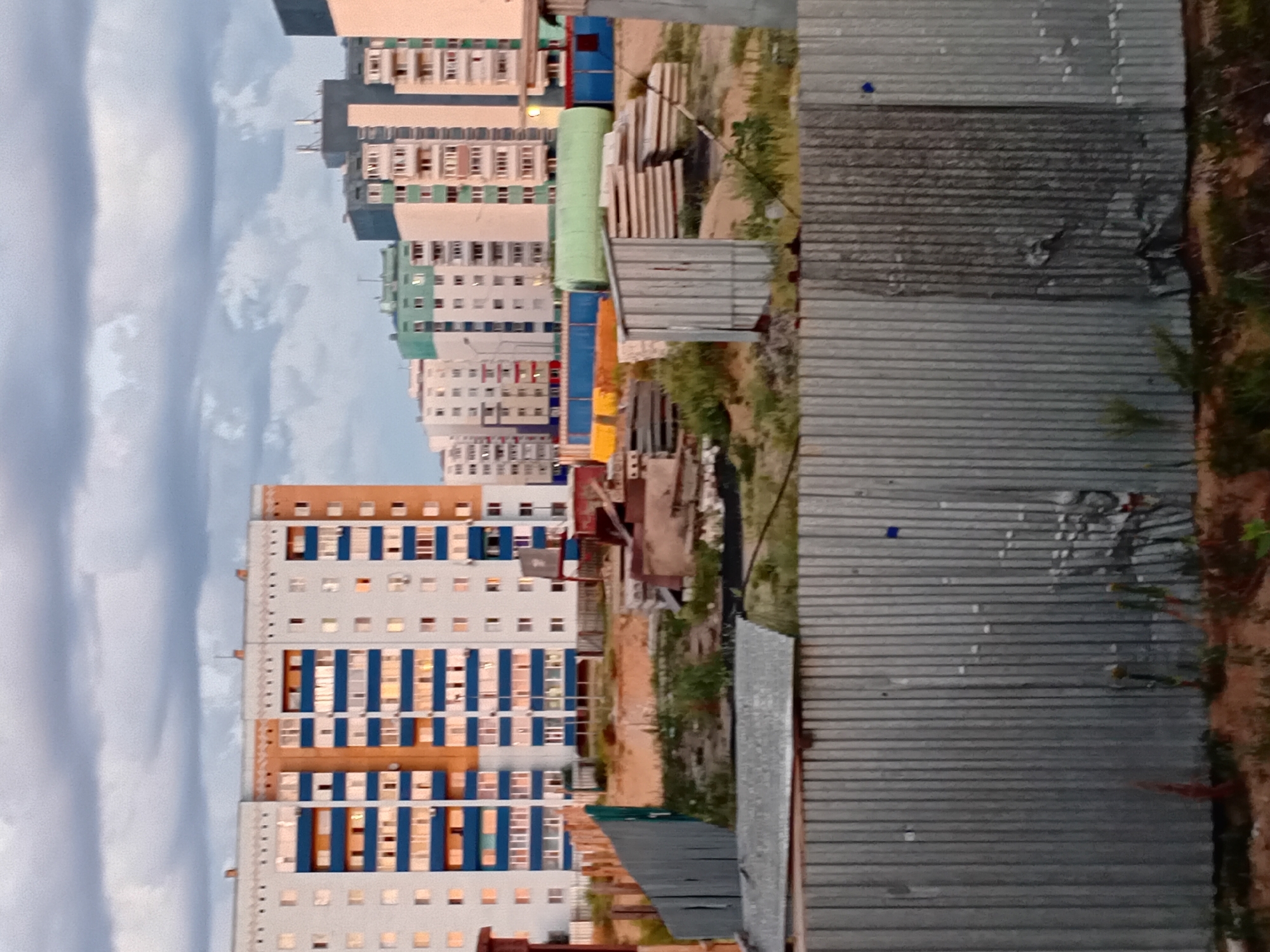 This image depicts a residential construction site in the city of Semey, Kazakhstan. In the background, several multi-story apartment buildings are visible, varying in architectural style and color. One prominent building features white and orange facades with blue balconies. In the foreground, a corrugated metal fence runs horizontally across the frame, partially obscuring the view of the construction area. The site itself is littered with construction materials, including stacks of concrete slabs, wooden planks, and a large green cylindrical object. The ground appears to be uneven and covered with dirt and sparse vegetation. The sky above is overcast with dramatic, dark clouds, suggesting either early morning or late evening light. No people are present in the scene.