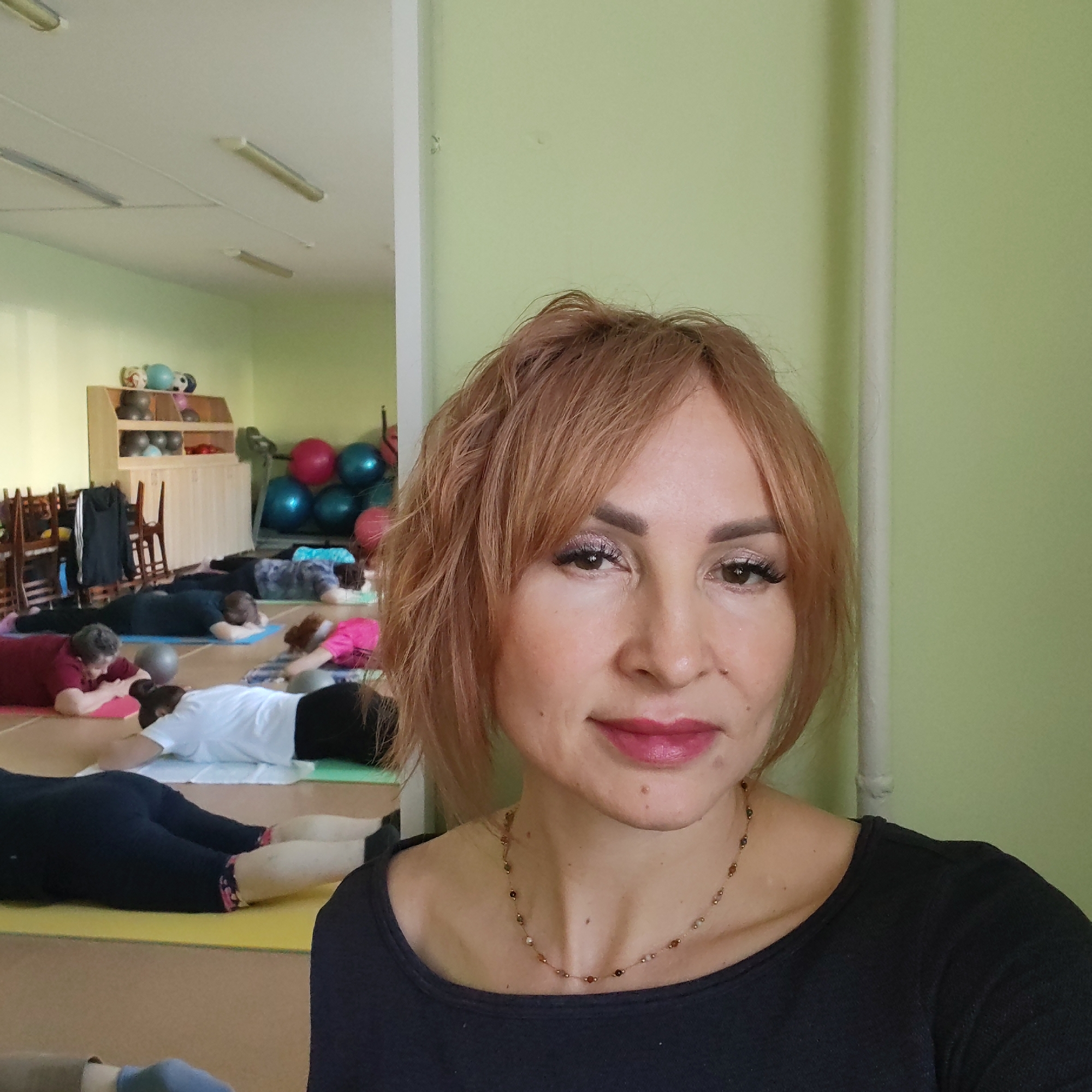 A middle-aged woman with red hair and fair skin is taking a selfie in a room filled with people doing yoga or pilates on mats. The woman is wearing a black top and a delicate necklace. In the background, there are several individuals lying on mats in what appears to be a yoga or exercise class. Some are using exercise balls. There is also shelving with various exercise equipment such as balls, and some chairs stacked in the corner. The room is painted with light green walls and fluorescent lights. The location is Chaikovsky, Russia, based on prior knowledge or context.