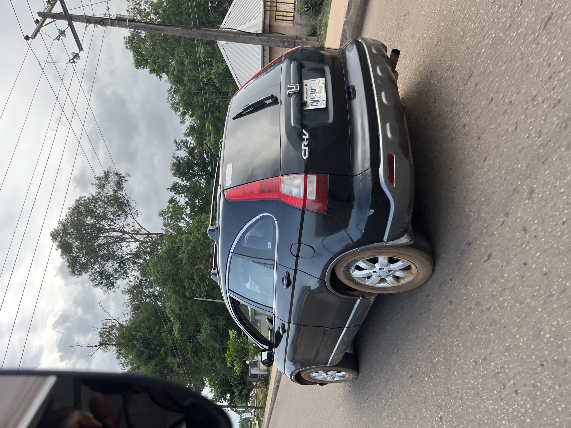 The image shows the rear passenger side of a black Honda CR-V driving on a paved road. The car has a roof rack and the license plate is visible, reading "GWA 104". There is a sticker on the rear windshield that says "JESUS". The car's wheels are covered in dirt. The environment appears to be a town or city in Nigeria, with trees and buildings visible in the background. A utility pole with power lines is also present. The sky is overcast. The scene suggests a typical day of transportation within Bukuru, Nigeria.