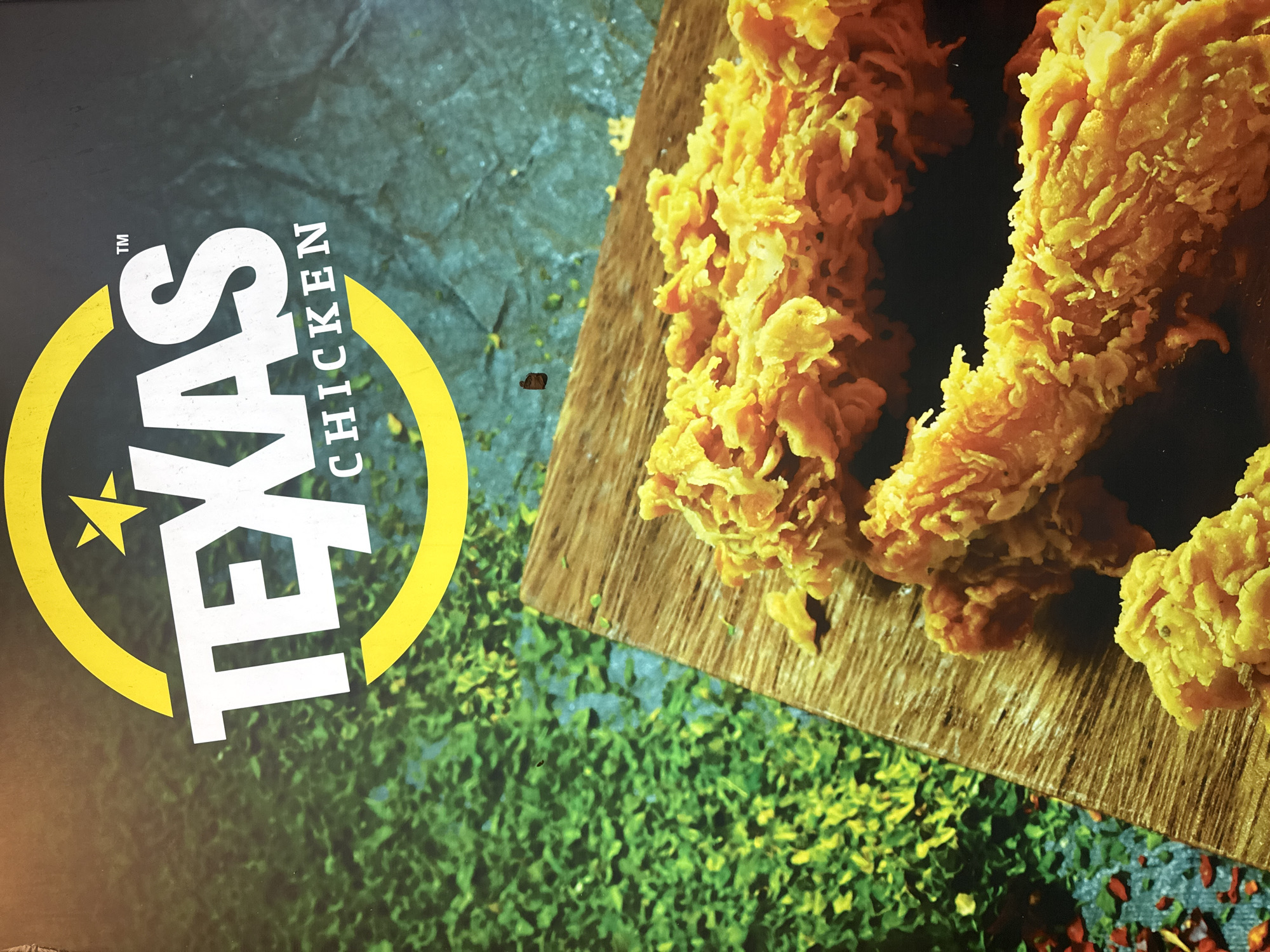 The image is a close-up photograph of a Texas Chicken restaurant advertisement. The logo, featuring a yellow circle with a white star and the words "TEXAS CHICKEN" in white text, is prominently displayed at the top of the image. Below the logo, a portion of a wooden board is visible, adorned with several pieces of fried chicken. The background is a blurred green and blue textured pattern, possibly representing foliage or a stylized landscape. The overall presentation is vibrant and appetizing, typical of fast-food advertising. There are no people or specific landmarks visible that would indicate a precise location within Nha Trang Ward, Vietnam.
