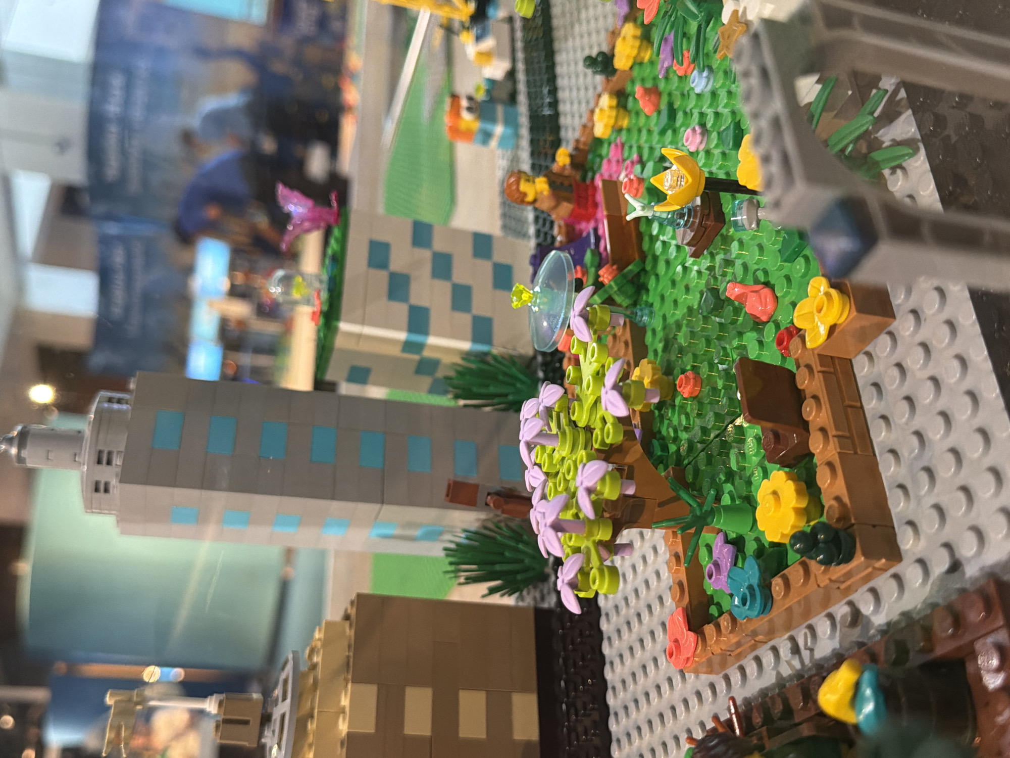 This is a close-up, eye-level, medium shot of a Lego model of a city. The scene is brightly lit with an artificial light source, and the focus is sharpest on the foreground and midground elements.
In the foreground, a detailed Lego garden is depicted. There is a small tree with purple flowers and green leaves. Various colorful Lego flower pieces, including yellow, pink, and light blue, are scattered throughout the green baseplates representing grass. A small brown planter box encloses a portion of the garden. A Lego figure with brown hair and a brown shirt is seated on a brown Lego boat-like structure within the garden. A clear, dome-shaped Lego piece with a yellow crystal on top sits near the figure.
Behind the garden, a tall, gray Lego skyscraper with light blue windows dominates the midground. To the left of the skyscraper, another Lego building is visible, constructed with brown and tan Lego bricks. In the background, two blurred figures are visible, one in a blue shirt and the other in a lighter colored shirt, standing near what appears to be a display. The background also shows a blurred, large, light blue cylindrical object. The overall impression is a meticulously crafted Lego urban environment. The location is not specified in the image.