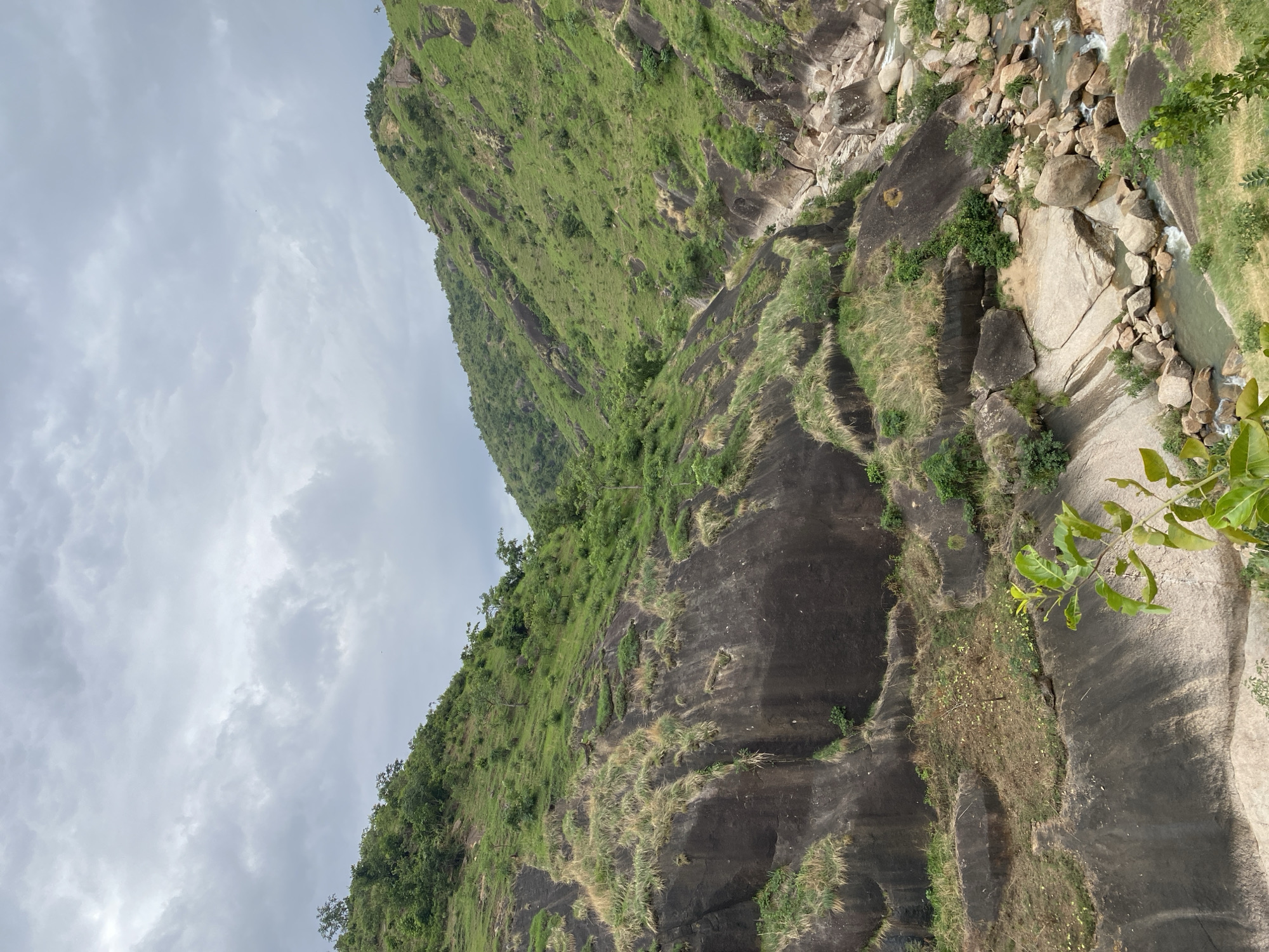 The image shows a rocky landscape with vegetation in Ganawuri, Nigeria. There is a steep rock face with patches of green vegetation and dry grass. To the right, a stream flows through rocks and boulders. The sky is cloudy, suggesting overcast weather. There are no people visible in the scene.