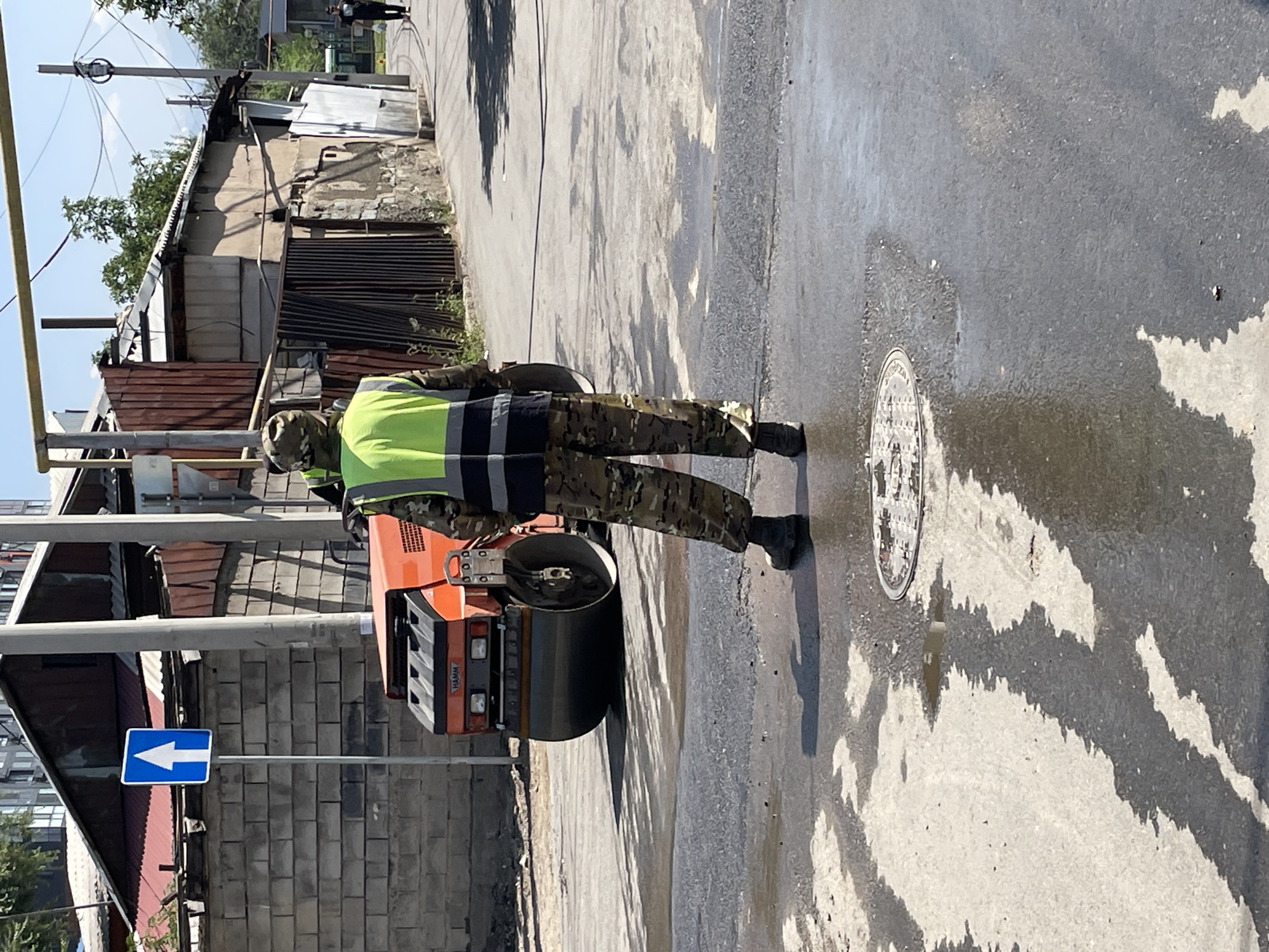 A male construction worker wearing a camouflage uniform and a bright yellow safety vest is standing on a wet asphalt road, looking towards a large orange steamroller. The steamroller is positioned behind him, and its front roller is visible. The scene appears to be a street under repair, with puddles reflecting the surroundings. In the background, there are several buildings, some appearing dilapidated, with corrugated metal roofs and brick walls. A blue and white directional sign indicating "go straight" is mounted on a pole to the left of the steamroller. Electrical wires are visible overhead, strung between poles. The sky is clear and blue. The overall impression is of ongoing roadwork in an urban or semi-urban area. The specific location is identified as the city of Almaty, Kazakhstan.