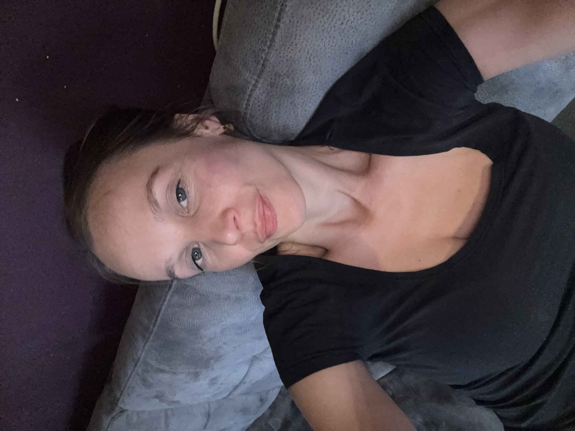 A medium shot captures a woman lying on a grey sofa against a dark purple wall. She is wearing a black t-shirt with a V-neck. Her head is tilted slightly to her left, and she is looking at the camera with a faint smile. Her hair is pulled back, and she is wearing a small hoop earring in her visible ear. The lighting appears to be dim, casting shadows on her face. The image does not provide any information about the specific location within Überherrn, Germany.