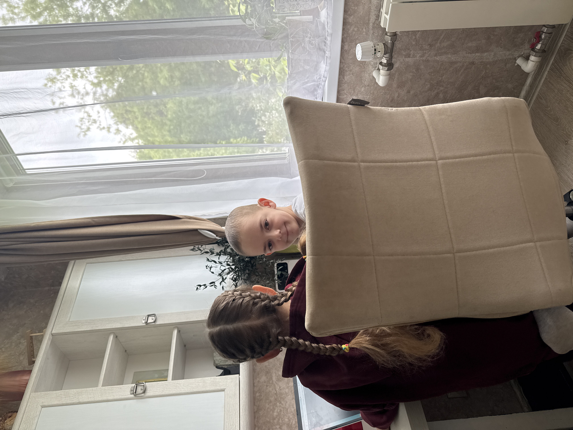 Two children are visible indoors. A younger child is peering from behind a large, beige cushion. An older child, wearing a maroon hoodie, is partially visible behind the cushion, with braided hair adorned with colorful elastics. A window with sheer curtains is behind the children, revealing a view of greenery. A radiator is visible on the wall beside the window. Part of a white cabinet or shelving unit is seen to the left of the children. The exact location is Razdolye, Russia.