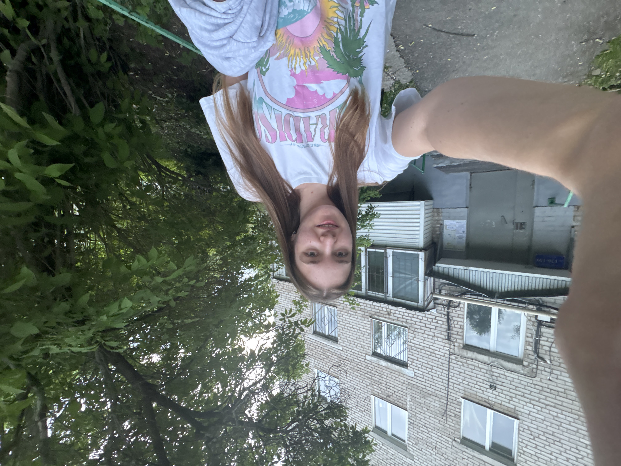 A young woman is taking a selfie outdoors, oriented in a disorienting perspective, in what is likely the city of Perm, Russia.
**People:**
* One young woman is the primary subject. She has long, straight brown hair and is wearing a white t-shirt with a colorful graphic design, as well as a grey sweatshirt draped over her shoulders.
**Environment:**
* The setting appears to be an urban outdoor space, possibly a courtyard or garden area beside a building.
* There is a tall brick building with multiple windows visible in the background. The architecture of the building is typical of urban Russian housing.
* Green trees and foliage surround the woman, filling the foreground on the left side of the frame.
**Objects:**
* A green clothesline is visible at the top left of the frame.
**Actions:**
* The woman is holding a camera (presumably a smartphone) to take a selfie.
**Inferences about location:**
* Based on the architecture of the building, and the general setting, it is likely that the photo was taken in the city of Perm, Russia.
* The picture is upside down.